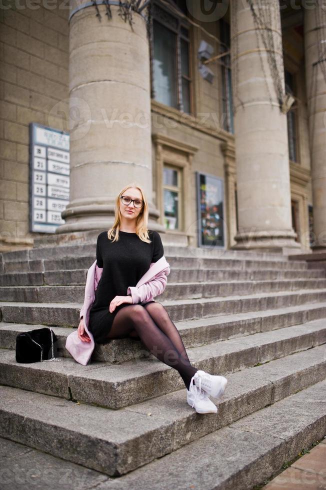 Blondes Mädchen mit Brille und rosa Mantel mit Handtasche sitzt auf der Treppe des Operngebäudes. foto