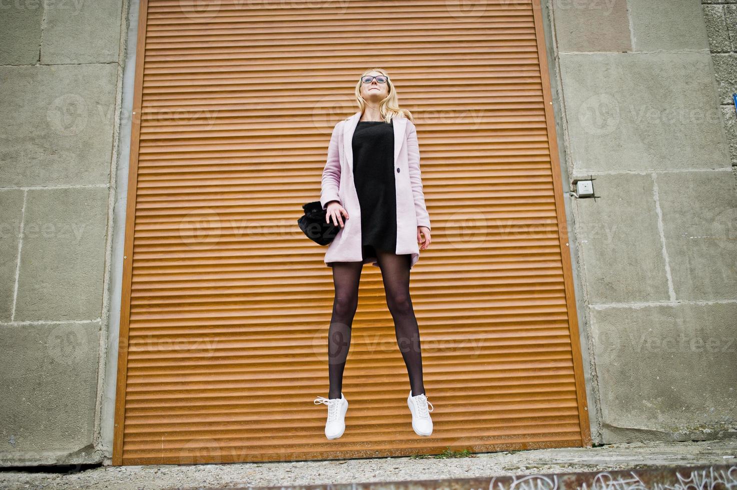 blondes mädchen mit brille und rosa mantel mit handtasche springen gegen fensterläden. foto