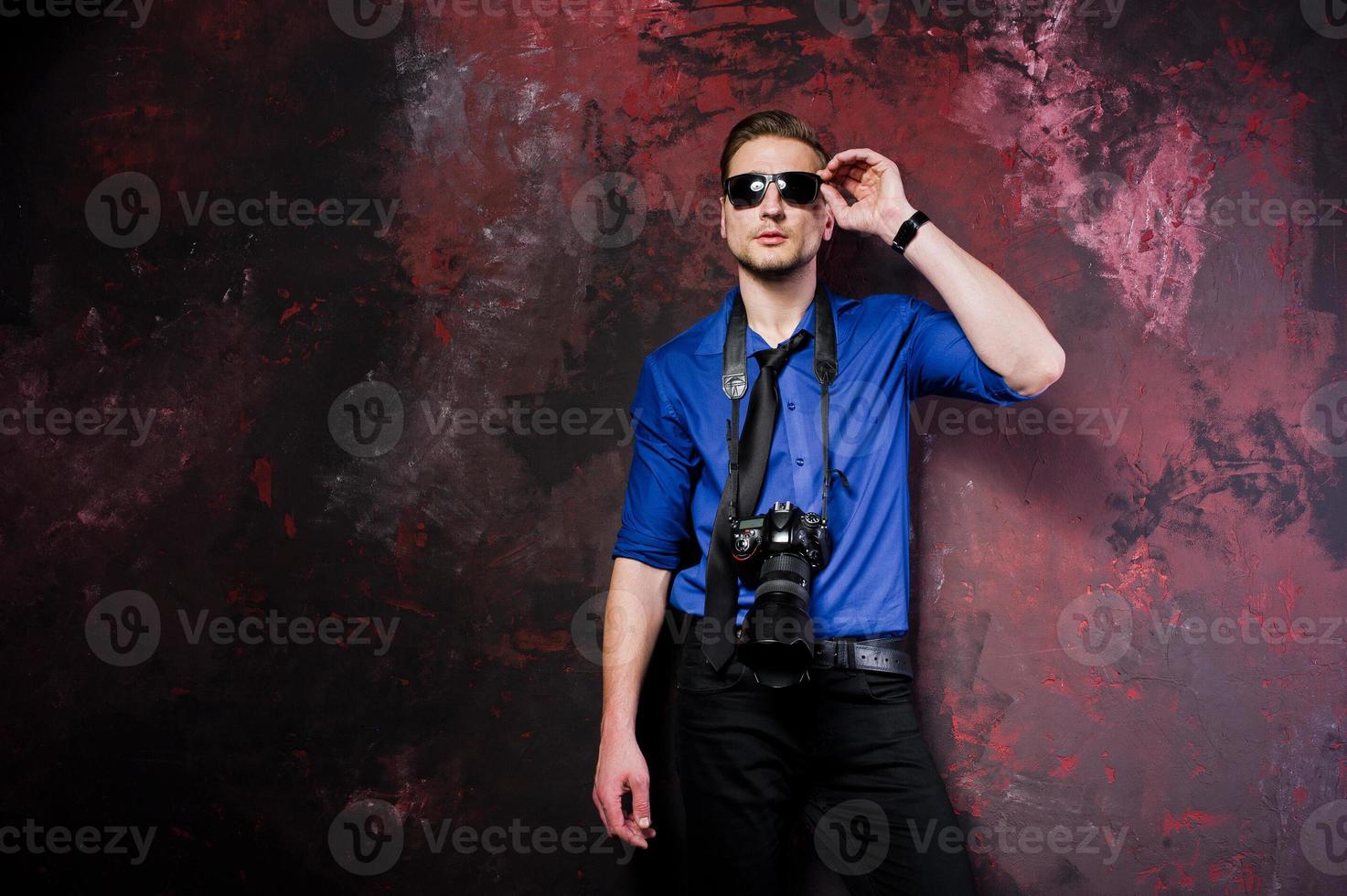 studioporträt eines stilvollen professionellen fotografenmannes mit kamera, tragen auf blauem hemd und krawatte, sonnenbrille. foto