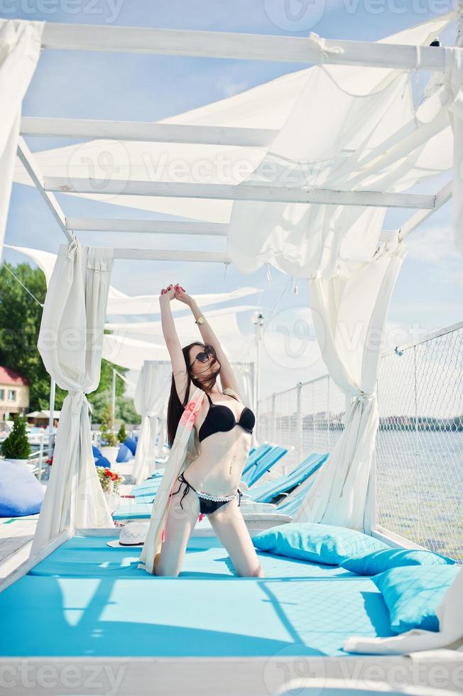Porträt eines erstaunlichen Mädchens im schwarzen Bikini-Badeanzug, das auf den Knien auf einer Liege am See posiert. foto