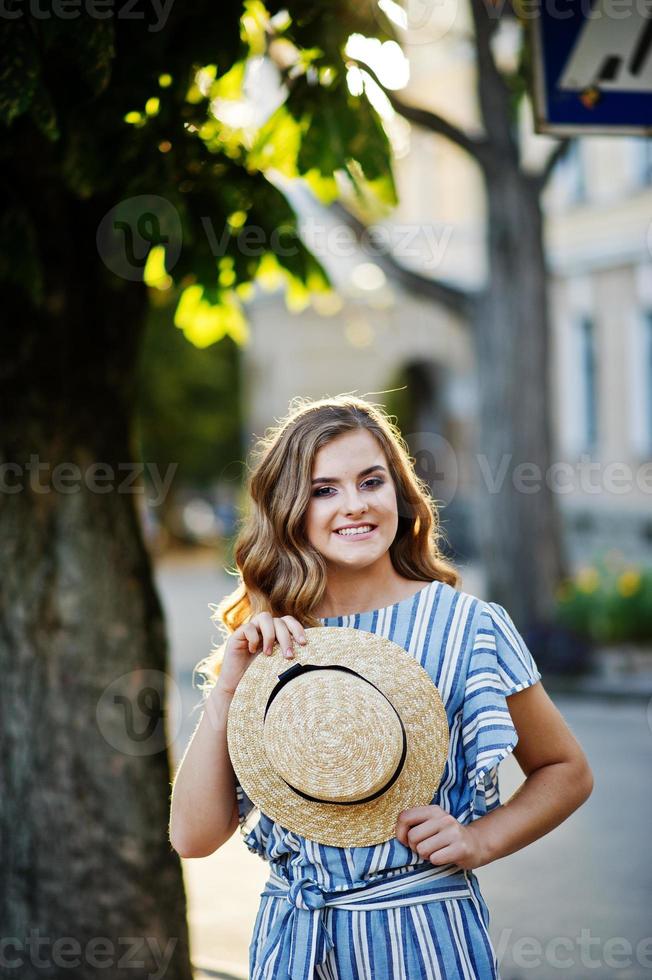 Porträt einer sehr attraktiven jungen Frau in gestreiftem Overall, die mit ihrem Hut auf einem Bürgersteig in einer Stadt mit Bäumen im Hintergrund posiert. foto