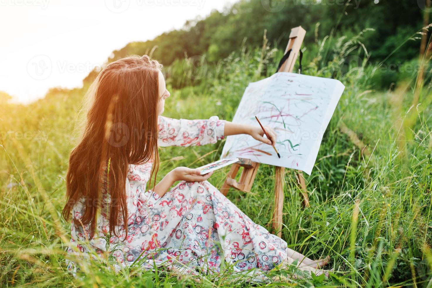 Porträt einer wunderschönen, glücklichen jungen Frau in schönem Kleid, die auf dem Gras sitzt und mit Wasserfarben auf Papier malt. foto