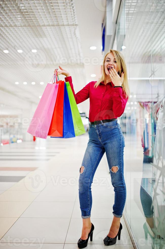 Porträt einer atemberaubenden jungen Frau in roter Bluse, zerrissenen Freizeitjeans und High Heels, die mit Einkaufstüten im Einkaufszentrum posiert. foto