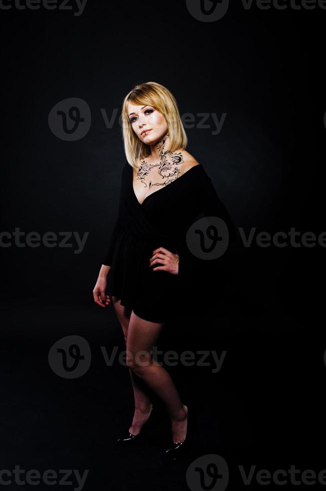 Studioporträt eines blonden Mädchens mit ursprünglichem Make-up am Hals, schwarzes Kleid auf dunklem Hintergrund. foto