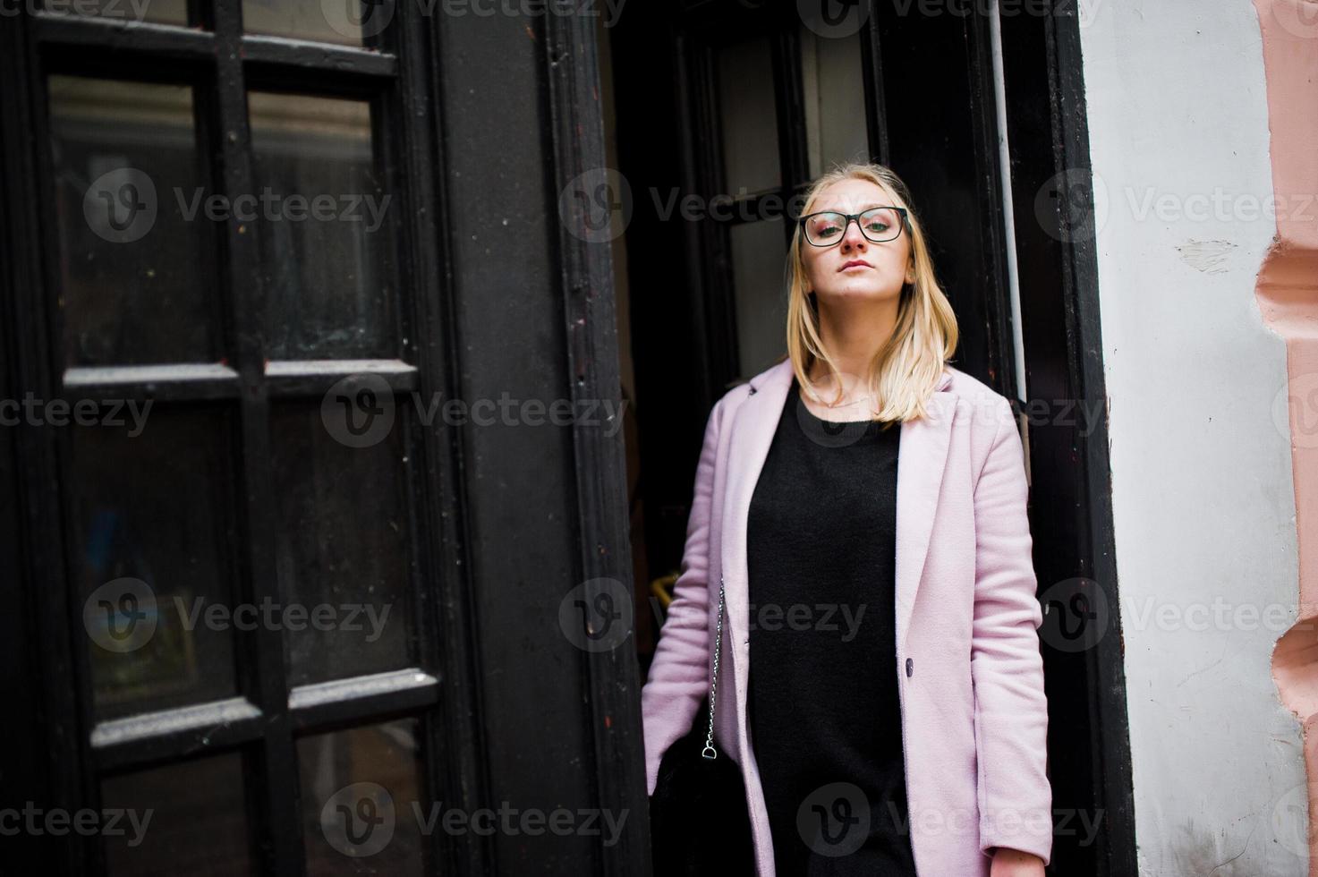 Blondes Mädchen mit Brille und rosa Mantel, schwarze Tunika posierte vor Holztüren. foto