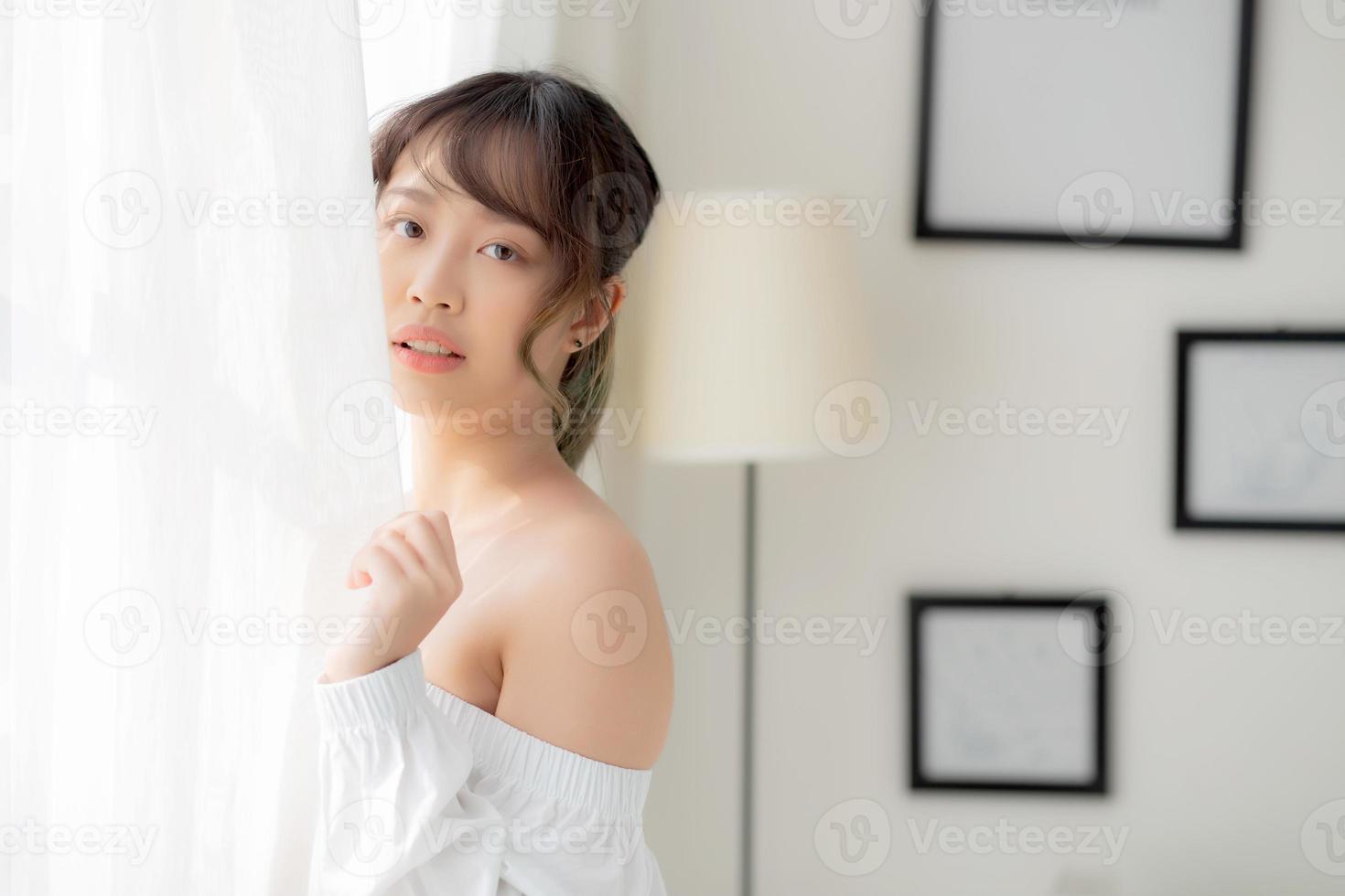 porträt einer schönen jungen asiatischen frau, die am fenster steht und lächelt, während sie morgens mit sonnenaufgang aufwacht, ein mädchen, das mit frischem und fröhlichem, lebensstil- und relaxkonzept zufrieden ist. foto