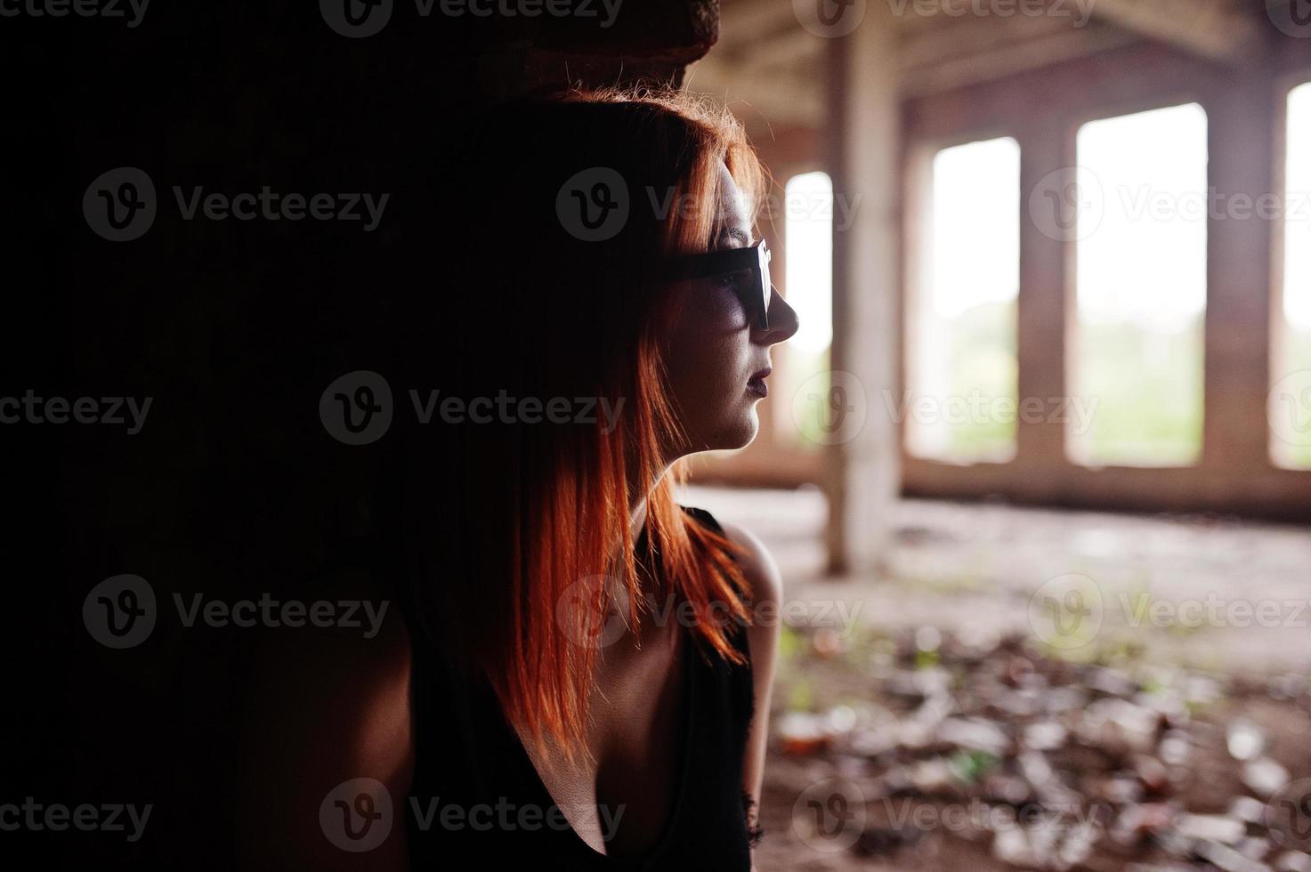 Rothaariges, stylisches Mädchen mit Sonnenbrille in Schwarz, vor verlassenem Ort mit Ziegelwänden. foto