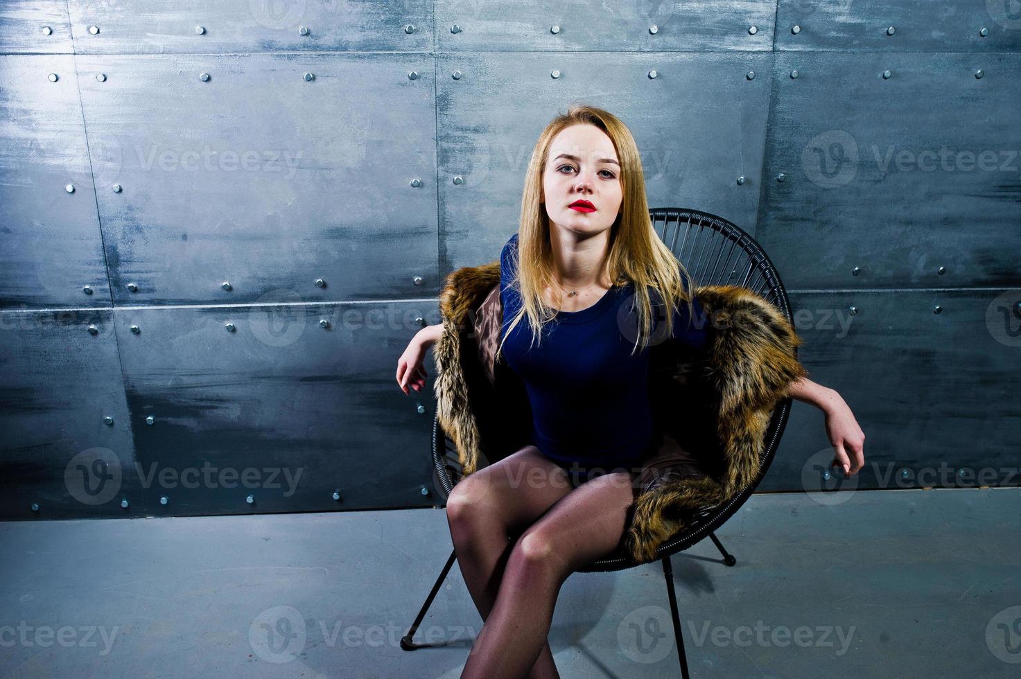 Elegante blonde Mädchen tragen einen Pelzmantel und ein Kombikleid, das auf einem Stuhl vor einer Stahlwand im Studio posiert. foto
