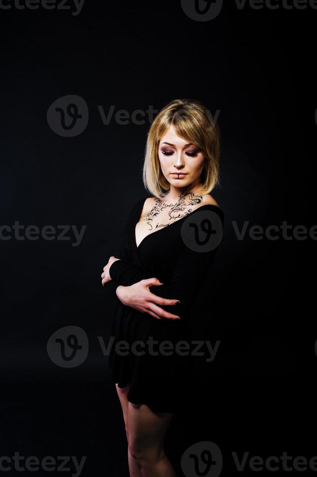 Studioporträt eines blonden Mädchens mit ursprünglichem Make-up am Hals, schwarzes Kleid auf dunklem Hintergrund. foto