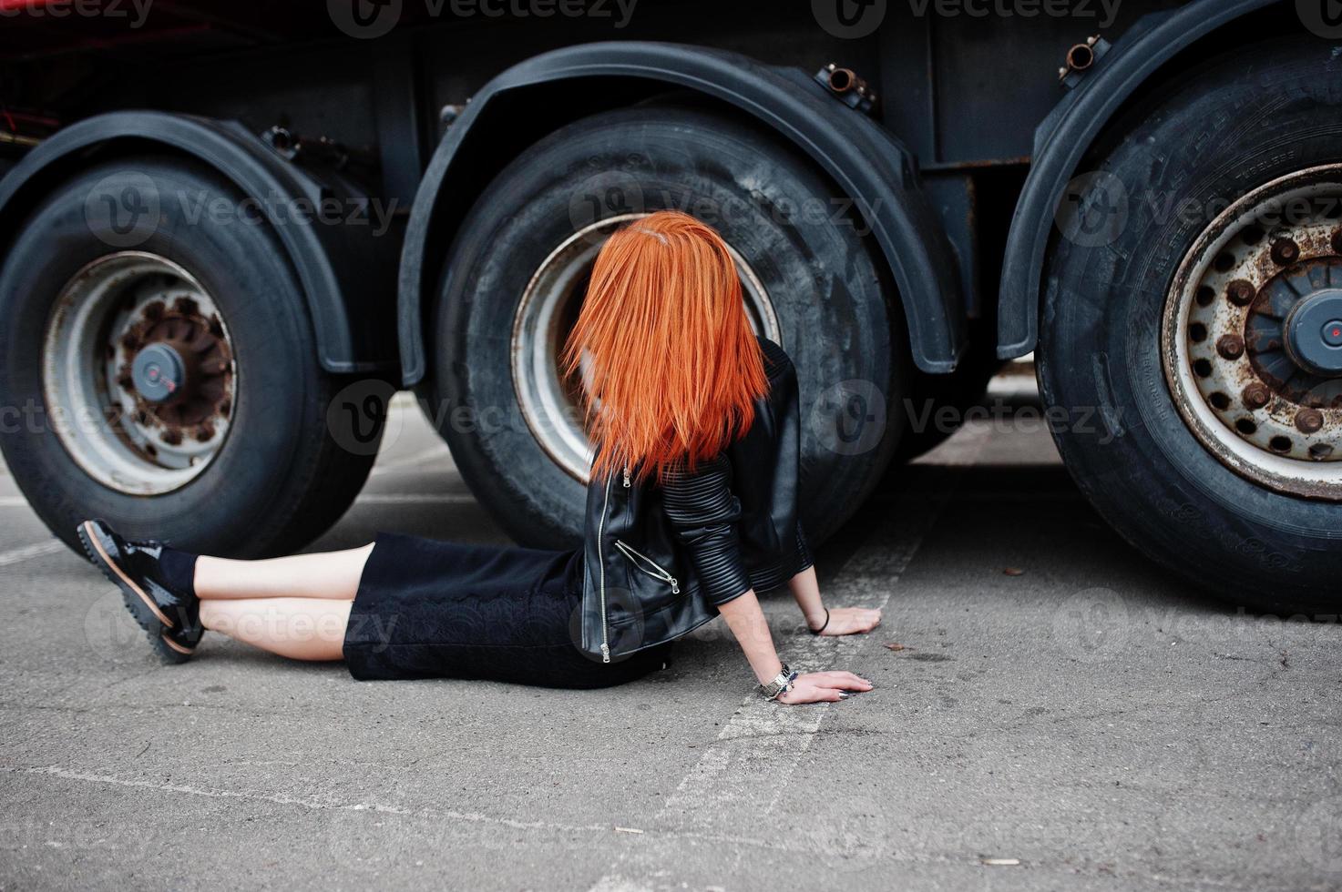 Rothaarige, stilvolle Mädchenkleidung in Schwarz, die an großen LKW-Rädern sitzt. foto