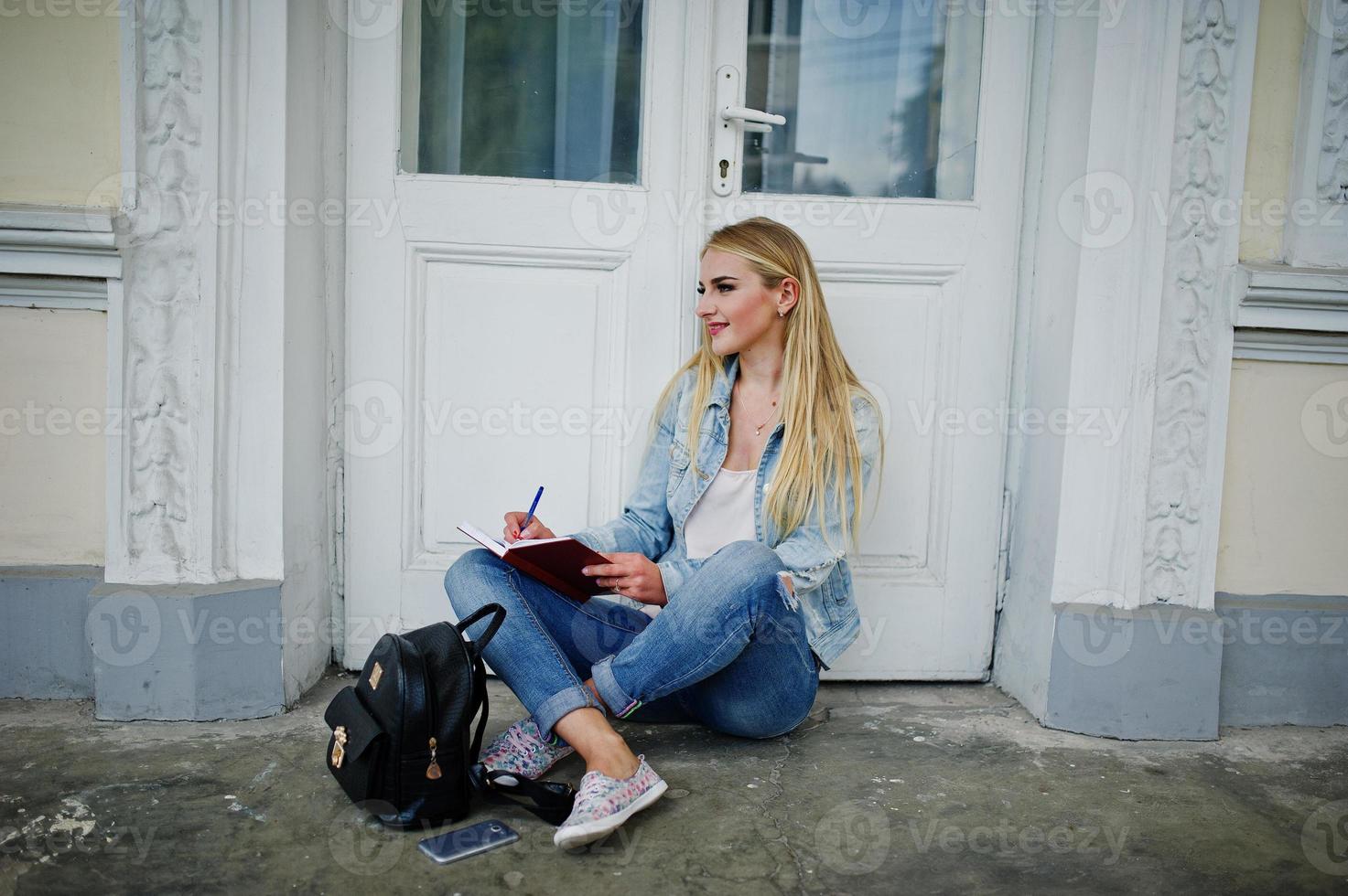 Blonde Mädchen tragen Jeans mit Rucksack, die mit Tagebuch gegen alte Tür gestellt wurden, und schreiben etwas. foto