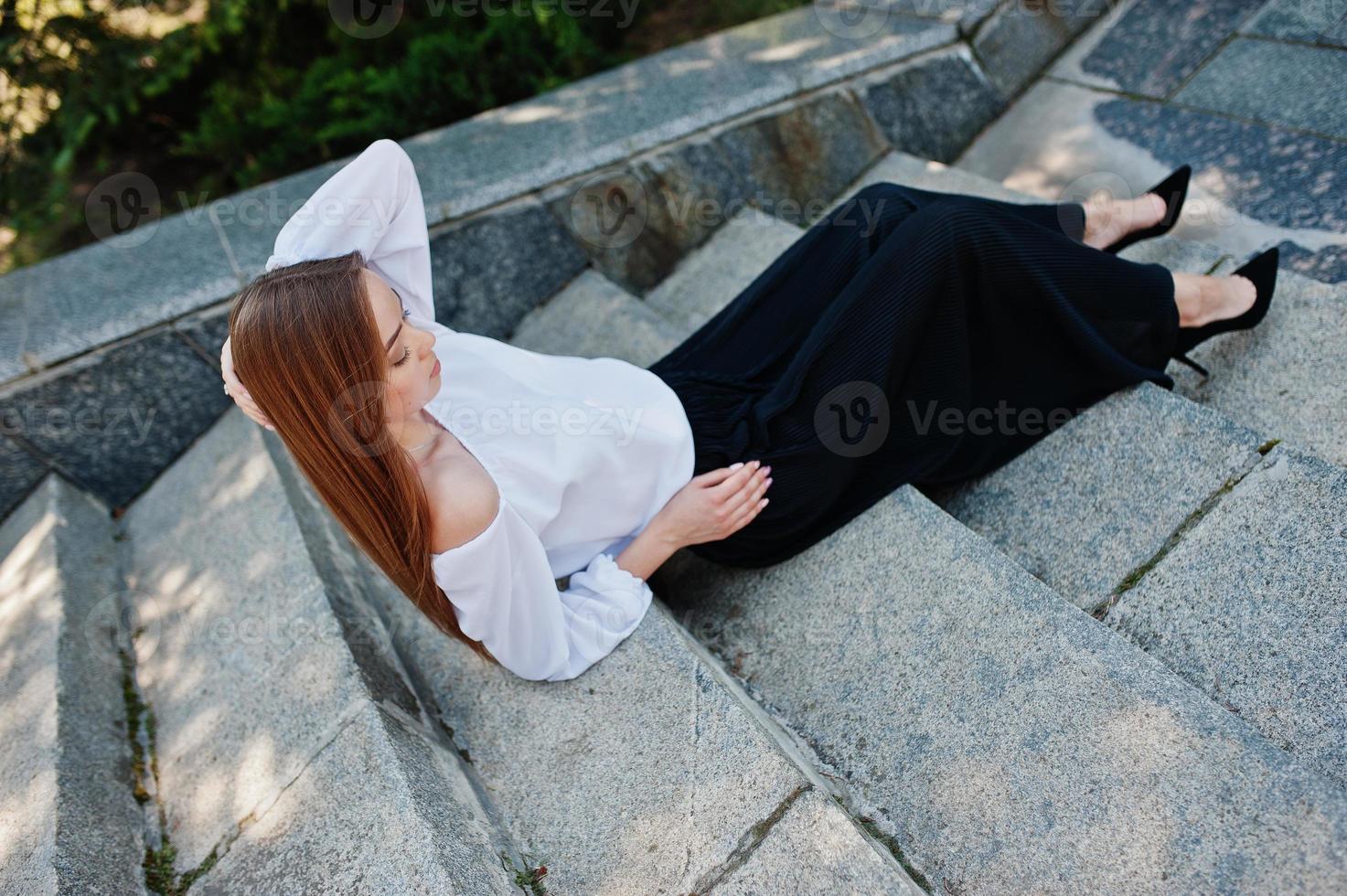 gut aussehende junge frau in weißer bluse, weite schwarze hose und schwarze klassische high heels, die auf treppen sitzen und posieren. foto