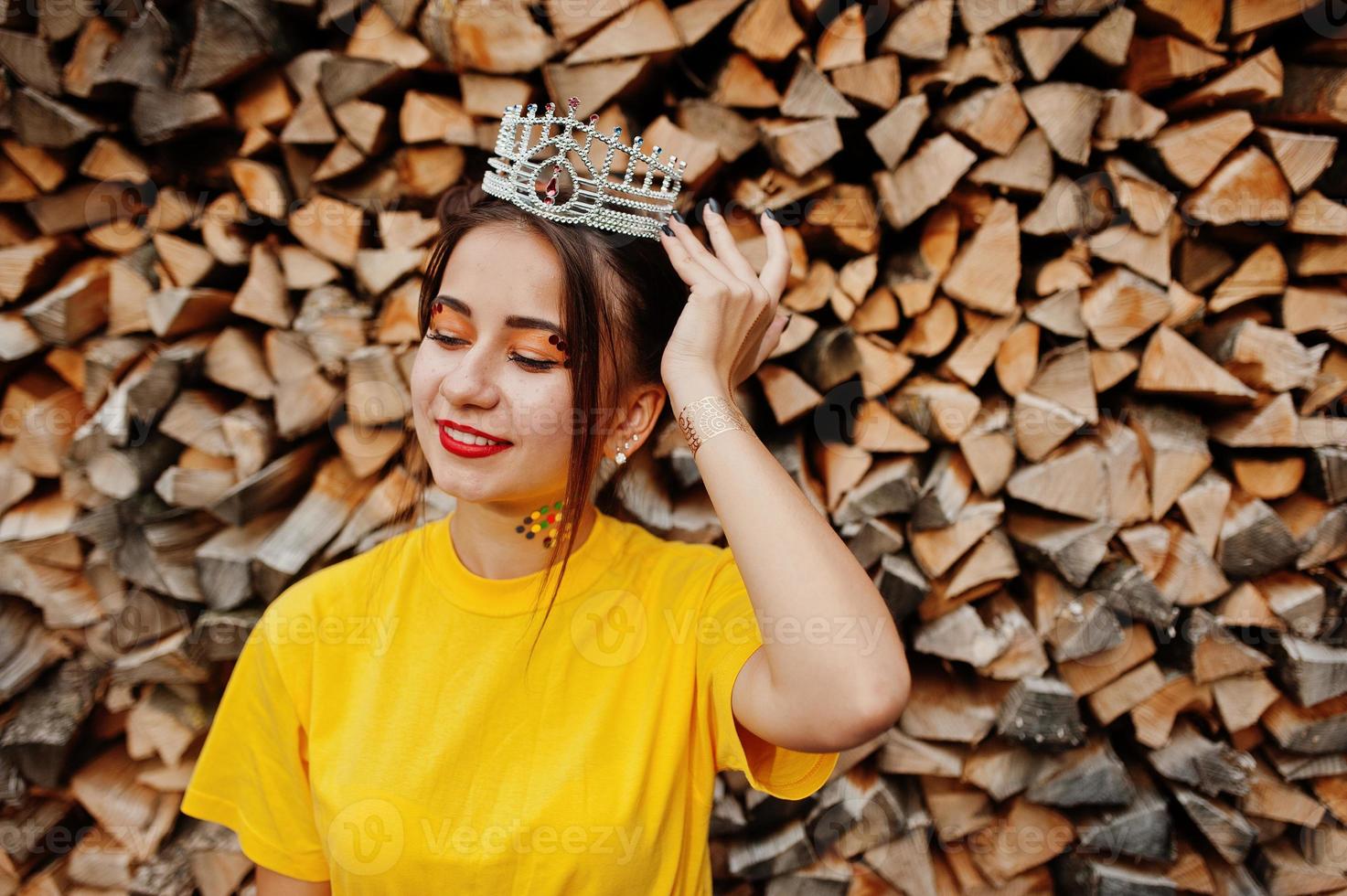 junges lustiges mädchen mit hellem make-up, wie märchenprinzessin, trägt gelbes hemd und krone gegen hölzernen hintergrund. foto