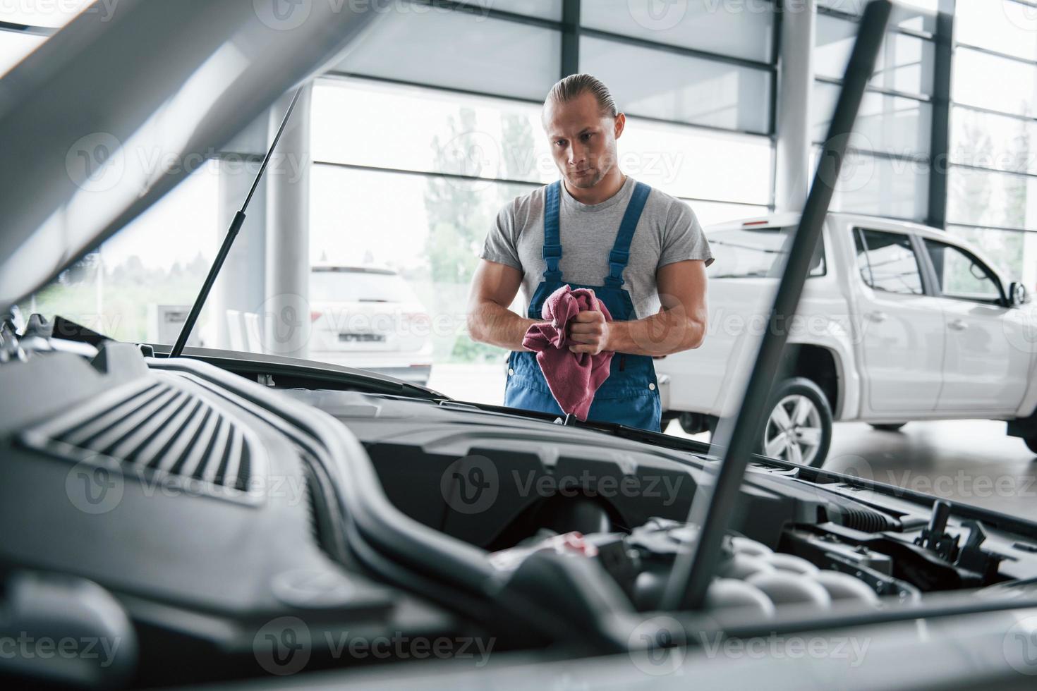 muss mal genauer hinschauen. Mann in blauer Uniform arbeitet mit kaputtem Auto. Reparaturen vornehmen foto