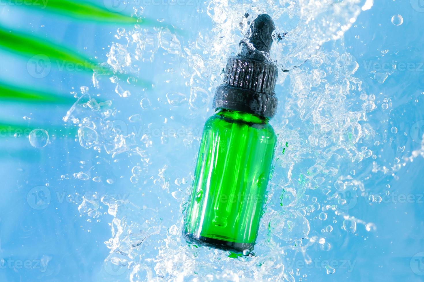 medizinisches hautpflegemodell, leere grüne glas-transparente serum-tropfflasche auf blauem wasserhintergrund mit wasser wird verteilt und spritzer verteilt. werbung schönheit kosmetisches produkt draufsicht. foto