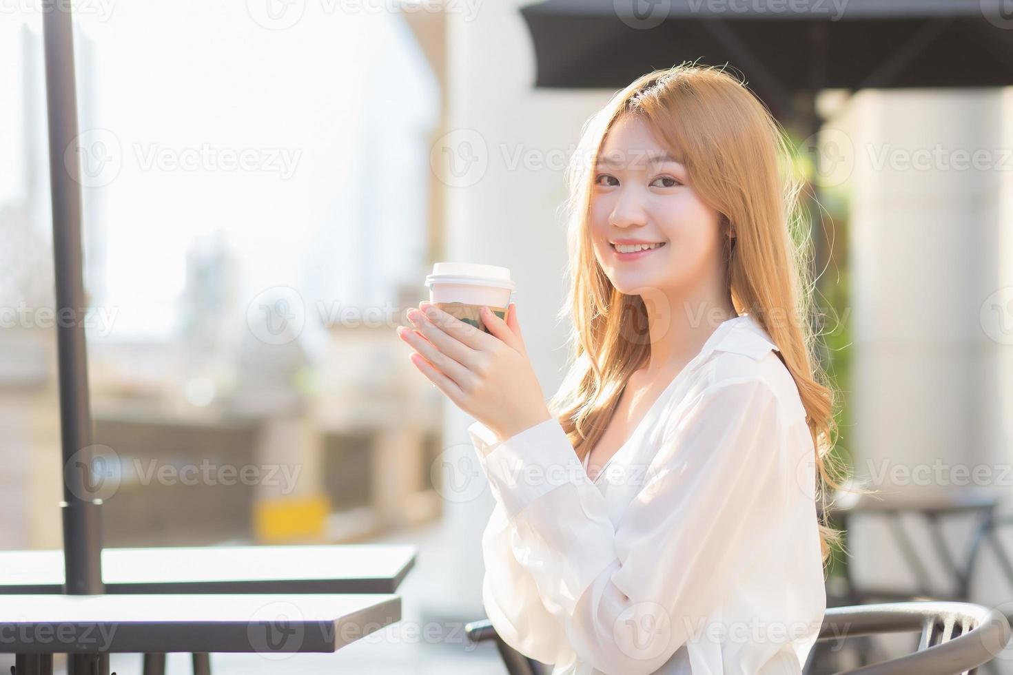 Schöne asiatische Frau, die einen Anzug mit bronzefarbenen Haaren trägt, sitzt lächelnd auf einem Stuhl im Café, während sie an einem sonnigen Morgen eine Kaffeetasse in der Hand hält. foto