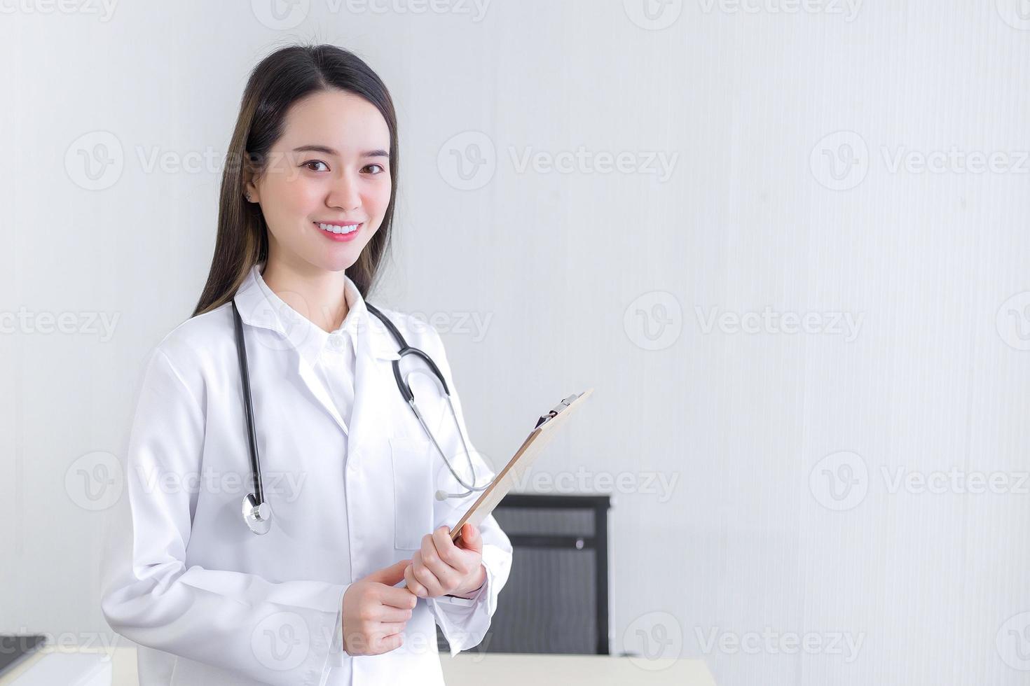Professionelle schöne junge Ärztin, die ein Dokument in der Zwischenablage hält und lächelnd in die Kamera schaut, während sie im Krankenhaus einen weißen Laborkittel und ein Stethoskop trägt. Gesundheitskonzept. foto
