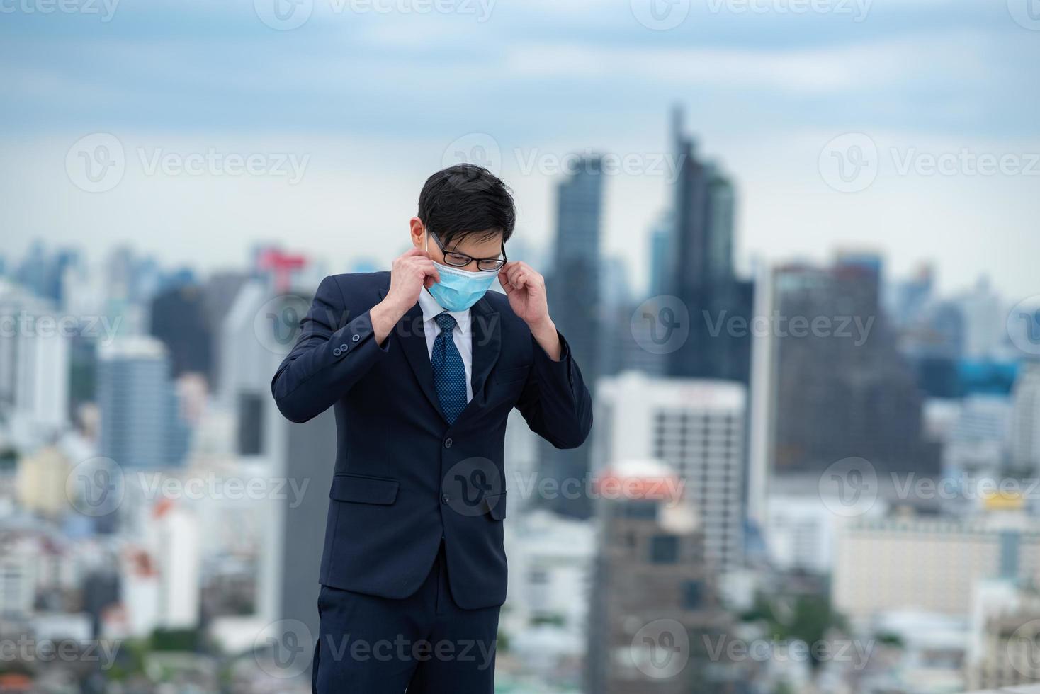 junger männlicher geschäftsmann, der covid-19-sicherheits-chirurgische maske trägt, während er auf der terrasse mit stadt und wolkenkratzern im hintergrund steht foto