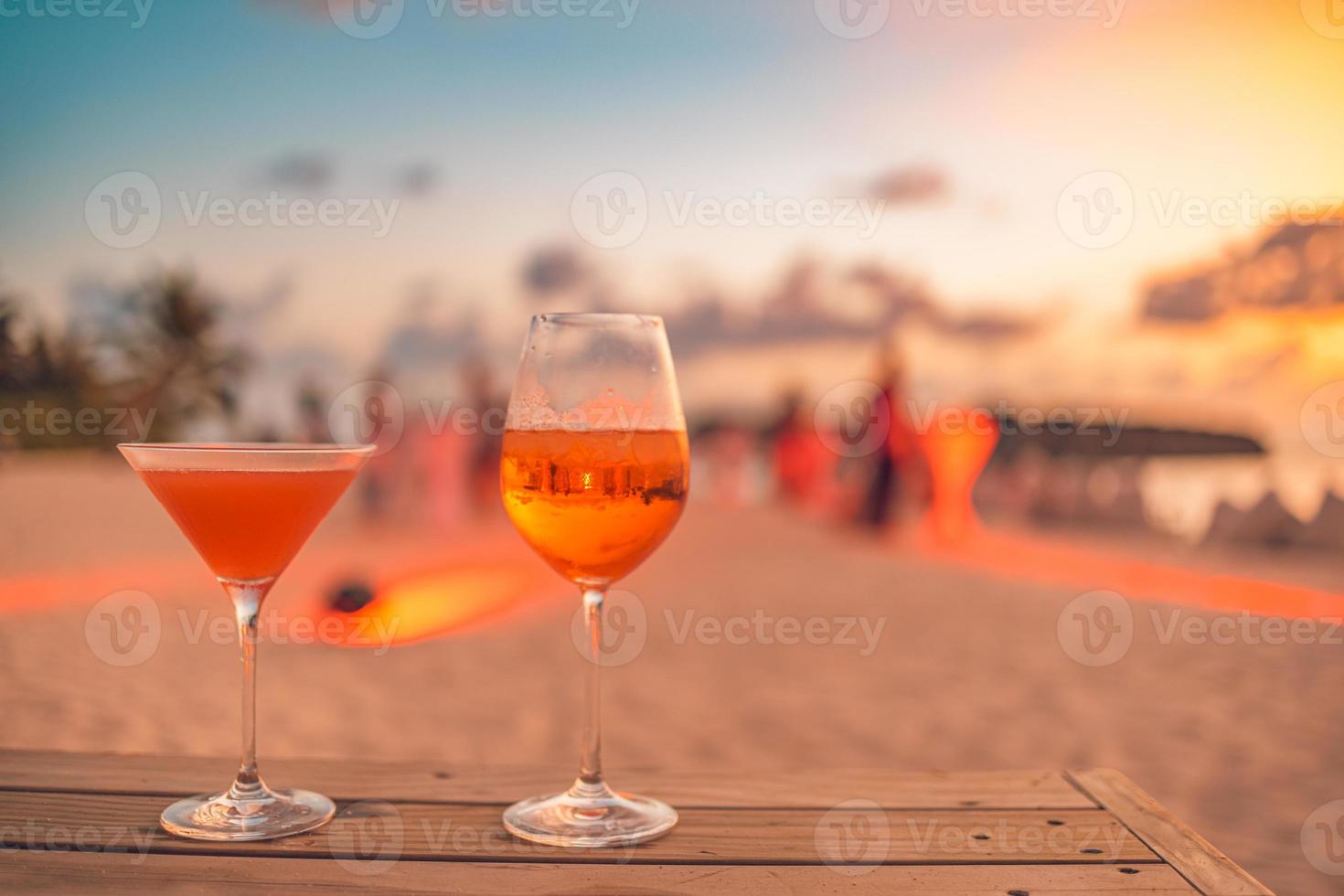 Zwei Cocktailgetränke mit unscharfen Strandparty-Leuten und buntem Sonnenuntergangshimmel im Hintergrund. luxuriöser Outdoor-Freizeit-Lifestyle, entspannende und romantische Farben, verschwommene Menschen, die an einem Sommerabend feiern foto