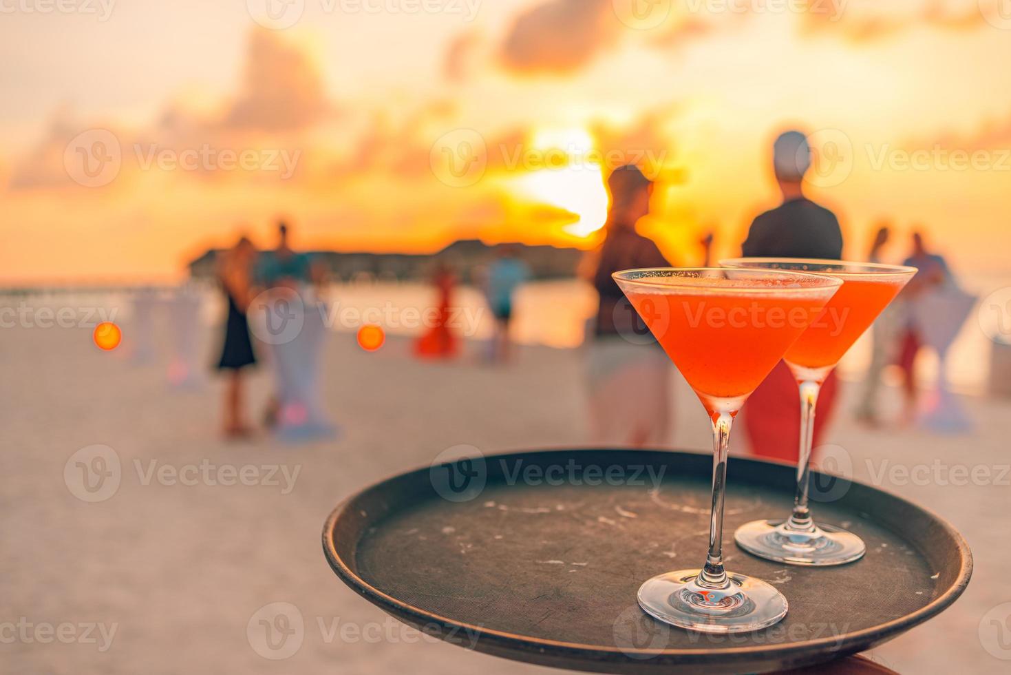 Zwei Cocktailgetränke mit unscharfen Strandparty-Leuten und buntem Sonnenuntergangshimmel im Hintergrund. luxuriöser Outdoor-Freizeit-Lifestyle, entspannende und romantische Farben, verschwommene Menschen, die an einem Sommerabend feiern foto