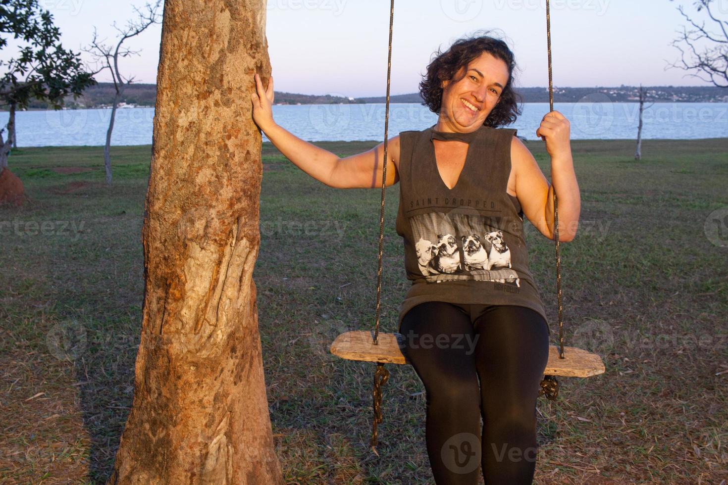 Reife Frau, die auf einer Schaukel in einem Park im Freien in der Nähe eines Sees schwingt foto
