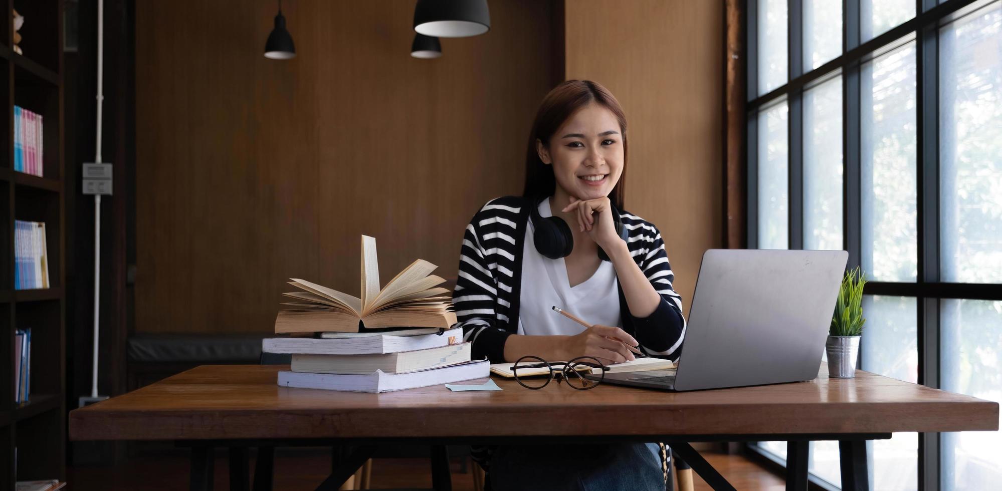 Schöne junge asiatische Studentin in der Bibliothek oder im Café, die ihre Hausaufgaben macht und einen Laptop benutzt. foto