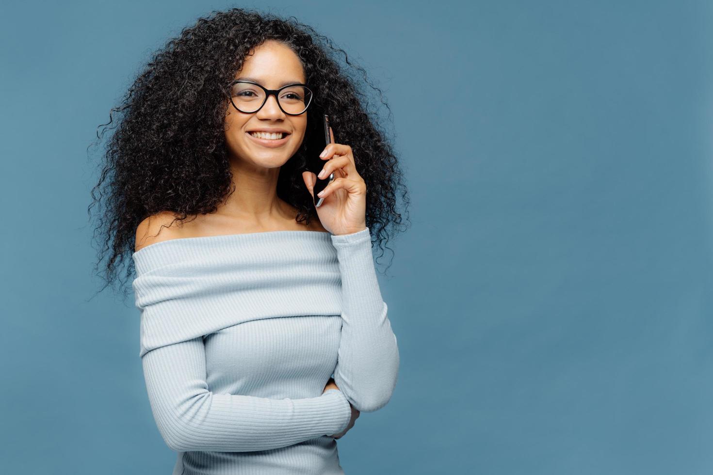 lächelnde begeisterte afroamerikanerin mit lockigem haar, spricht über smartphone, bespricht einige angenehme momente nach der party, schaut glücklich in die ferne, trägt brille und pullover, isoliert auf blauer wand foto