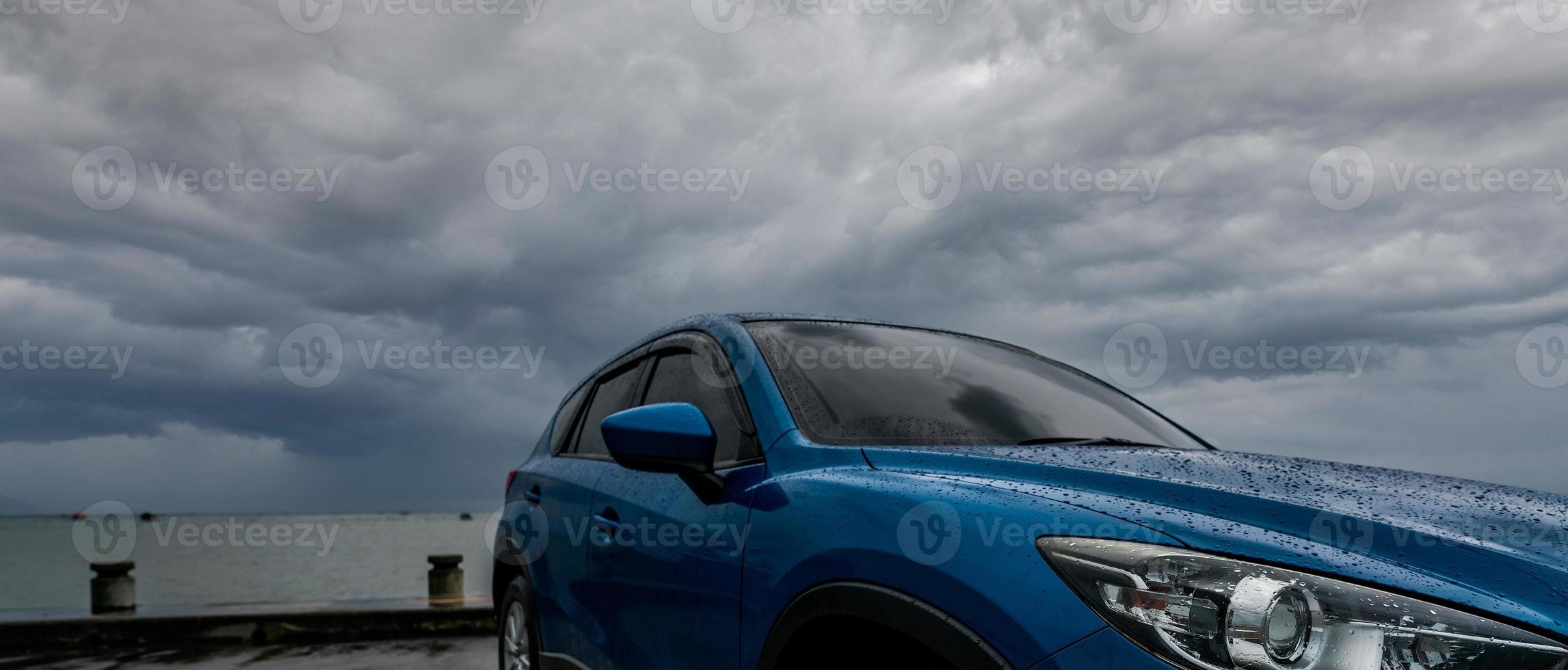 Blaues SUV-Auto mit Wassertropfen, das auf dem Parkplatz in der Nähe des Meeresstrandes gegen stürmischen und bewölkten Himmel geparkt ist. Vorderansicht des neuen Luxus-SUV-Autos mit Sportdesign. Mietwagen für Roadtrip. Autofahren an regnerischen Tagen. foto