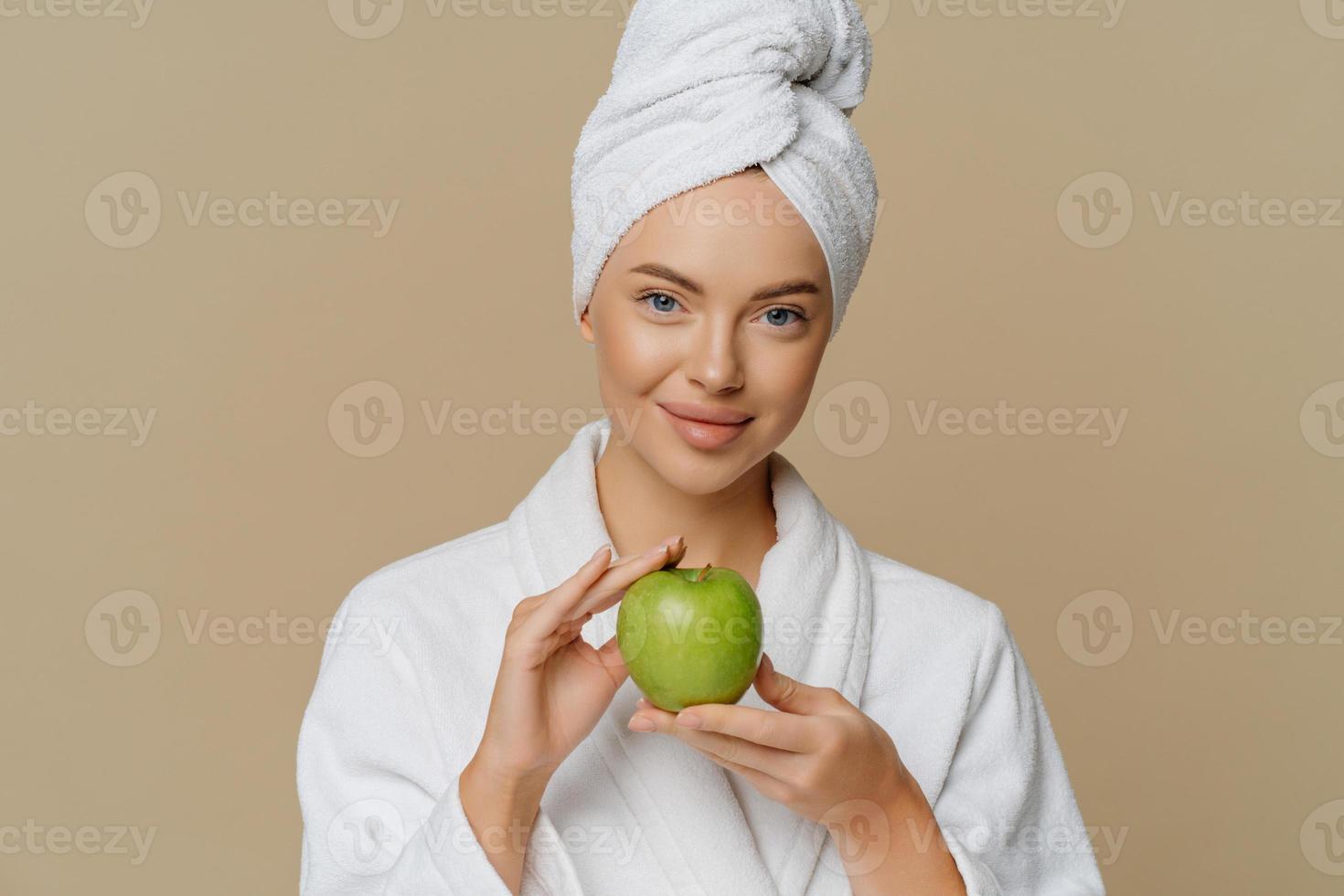 Schöne junge Frau hat die Haut erfrischt, nachdem sie ein Bad genommen hat, gekleidet in einen Bademantel, eingewickeltes Handtuch auf dem Kopf, hält einen grünen Apfel, der viele Vitamine enthält, die über braunem Hintergrund isoliert sind. Hygienekonzept foto