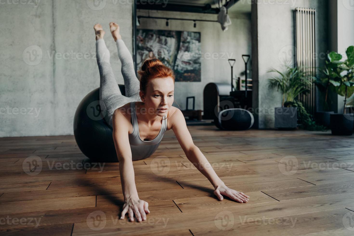 athletische schlanke dame mit roten haaren, die während des pilates-trainings streching-übungen auf einem großen silbernen fitball durchführt foto