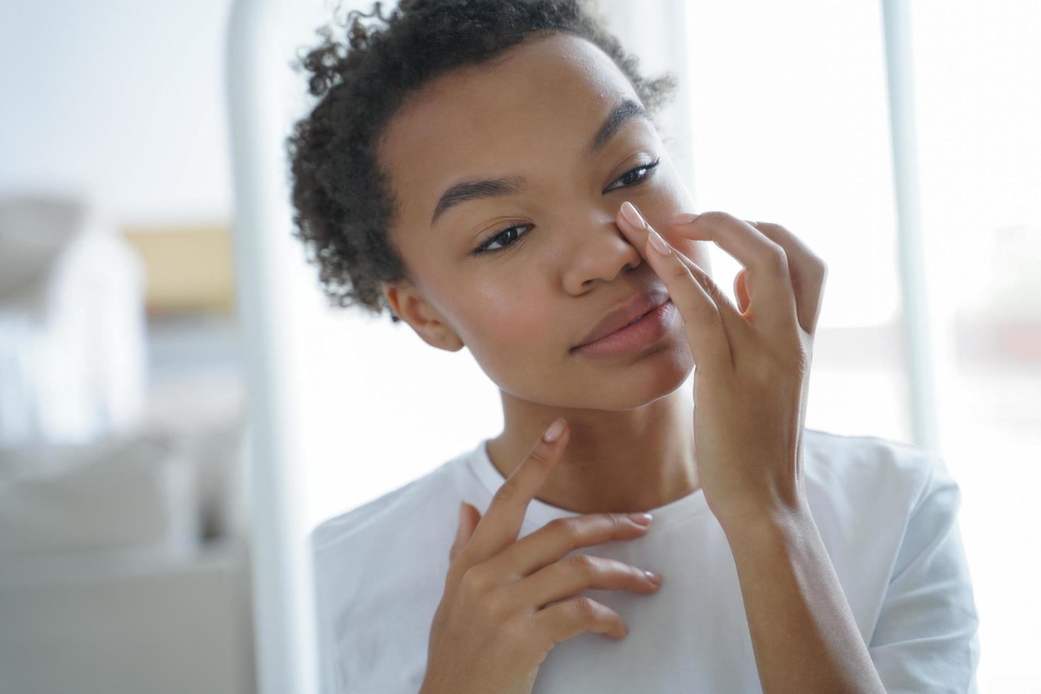 attraktives afro-mädchen trägt creme auf, die ihr gesicht berührt. Spiegelbild der jungen lateinischen Frau. foto