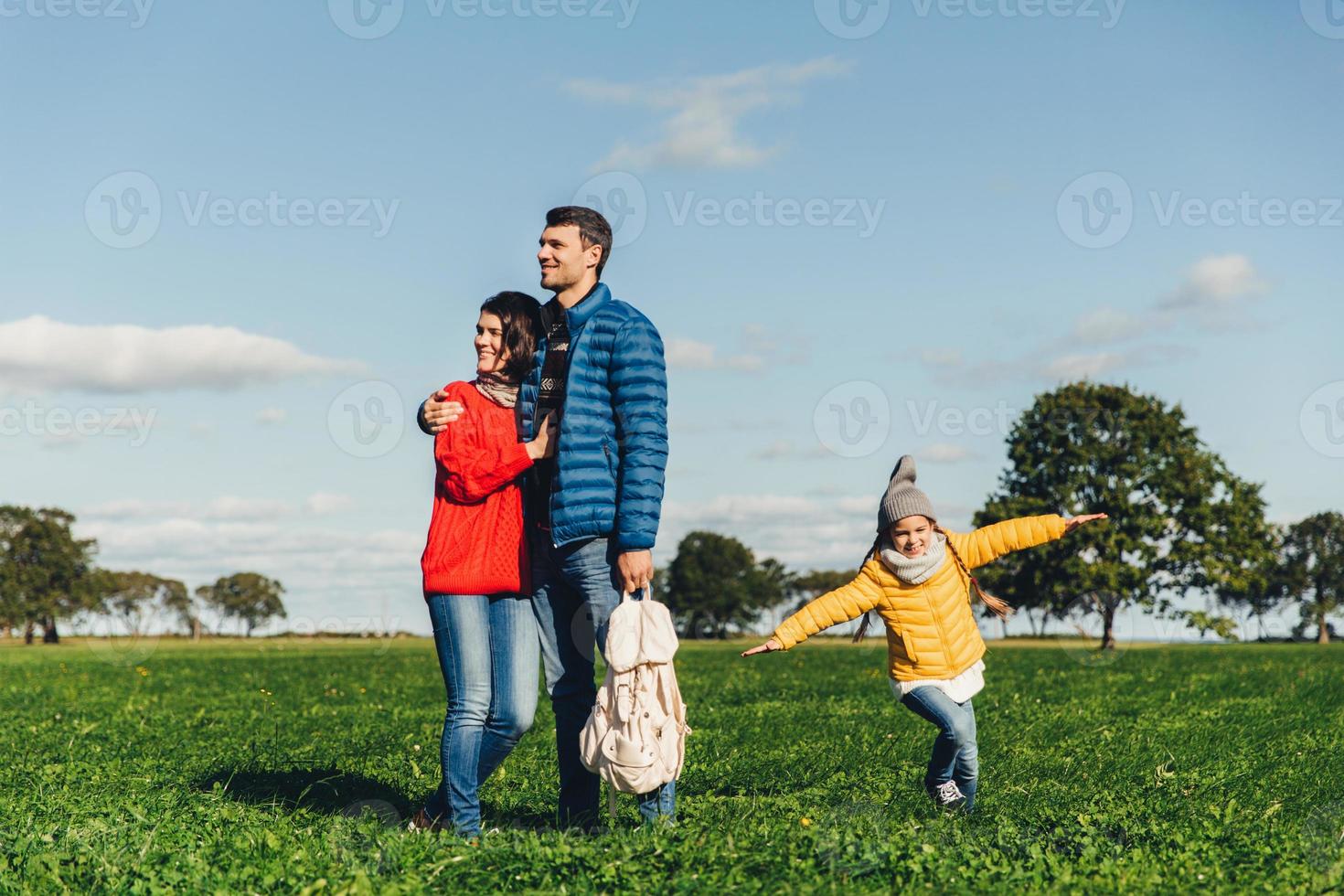 junge verliebte paare stehen auf grünem gras, umarmen sich und schauen nachdenklich. verspieltes kleines kind genießt freiheit, rennt auf feld, spielt in der nähe seiner eltern, hat gute laune und erholung. Mensch und Natur foto