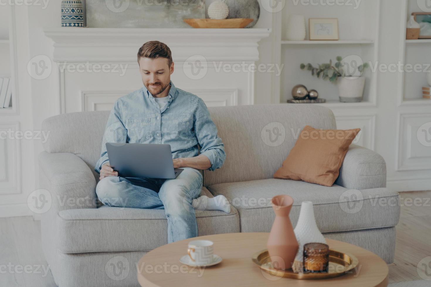 junger büroangestellter, der von zu hause aus arbeitet, während er mit gekreuztem bein auf der couch sitzt foto