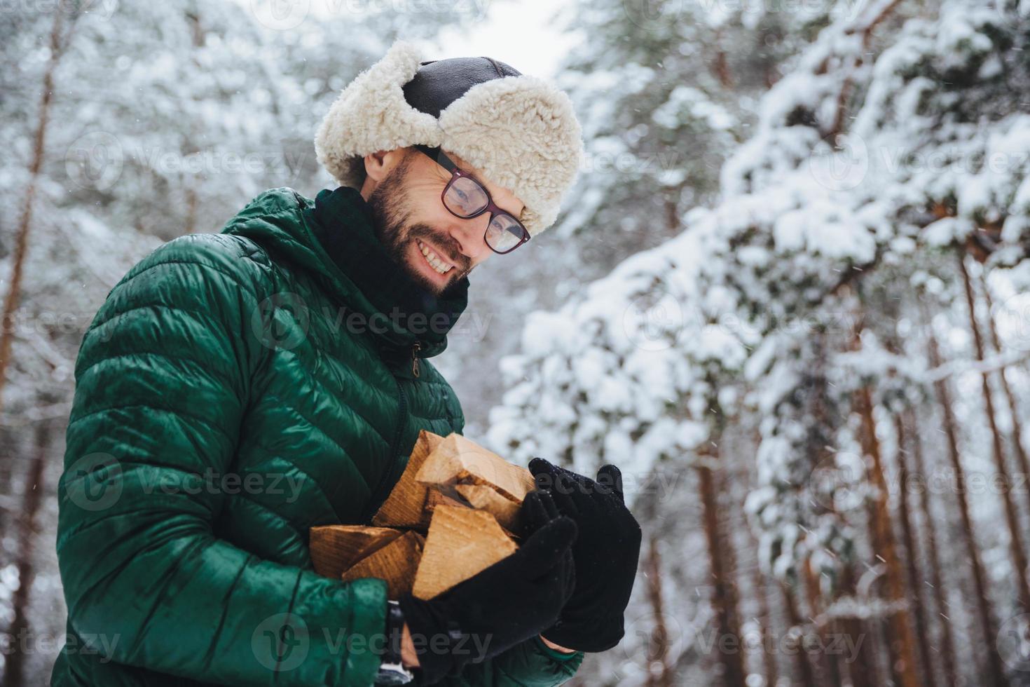 zufriedener mann mit brille schaut glücklich auf brennholz, das er im winterwald schlägt, um feuer zu machen, trägt grünen anorak, posiert vor weißem winterwald. menschen, lebensstil, erholungskonzept foto