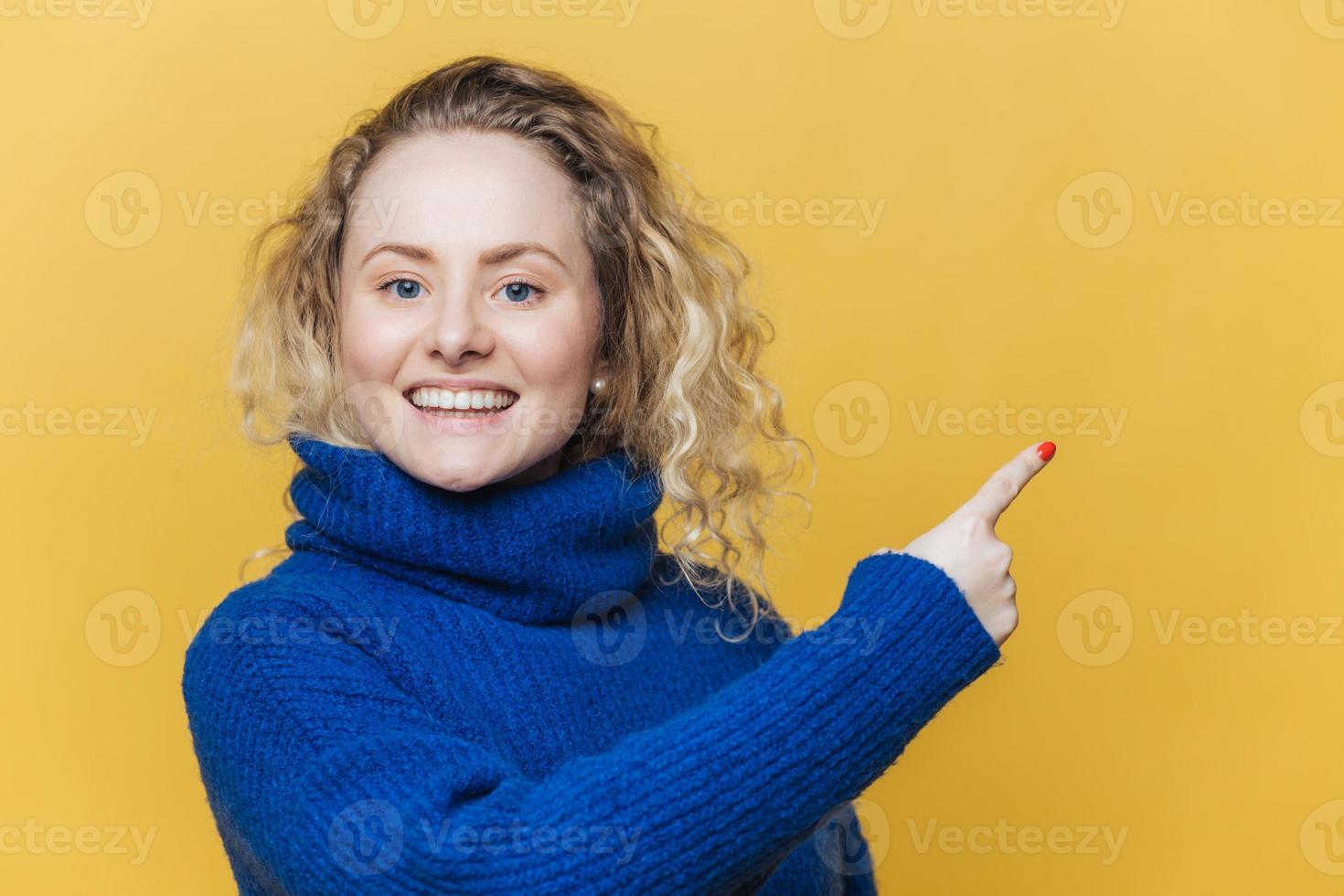 glückliche schöne blonde junge frau hat erfreuten ausdruck, trägt lässigen blauen pullover, zeigt auf leeren kopierraum auf gelber leerer studiowand an, wirbt mit großer vergnügen für etwas. Werbung foto