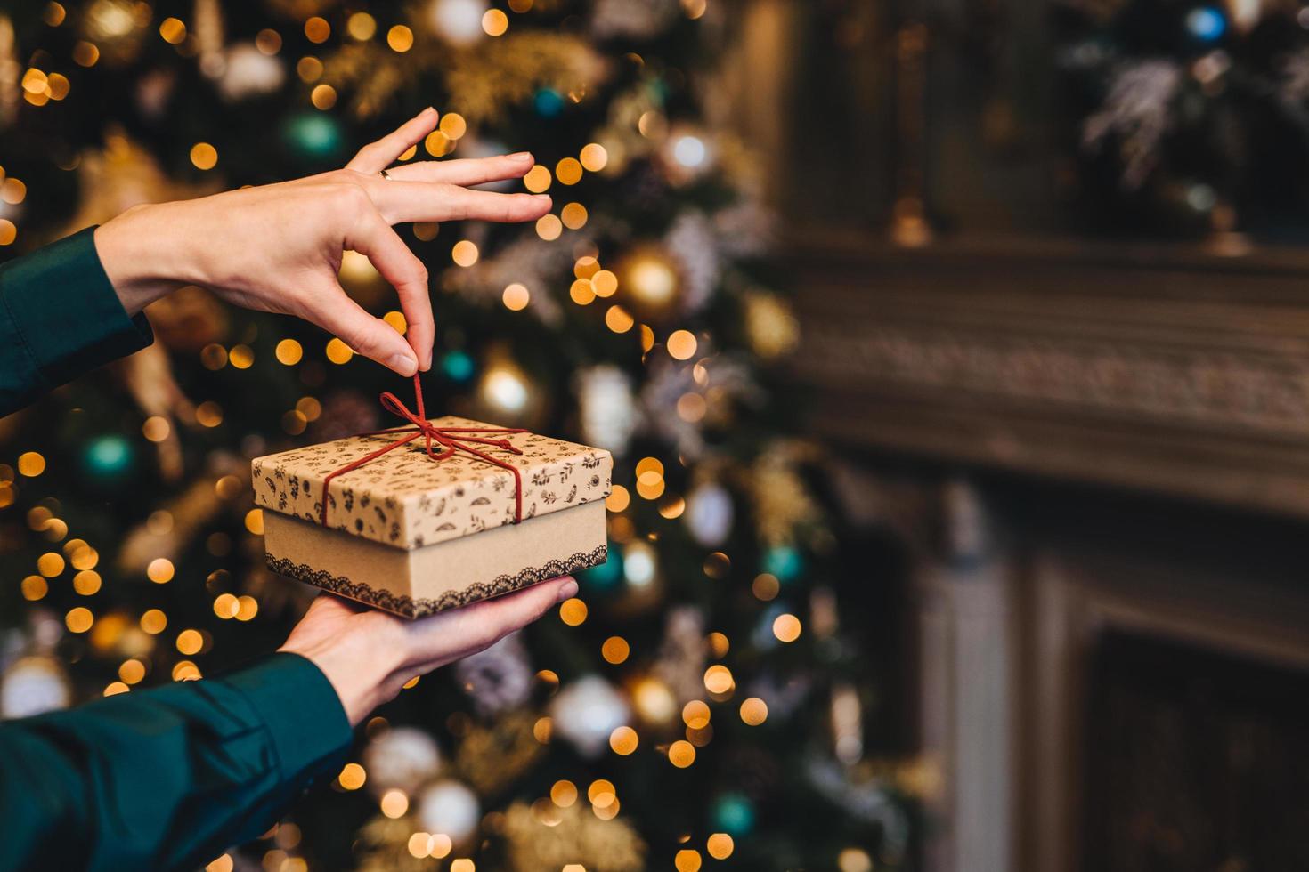 überraschung und angenehme momente konzept. frau verpackt neujahrsgeschenk als steht im wohnzimmer in der nähe eines schön dekorierten tannenbaums. feiertage, x mas und feierkonzept. foto