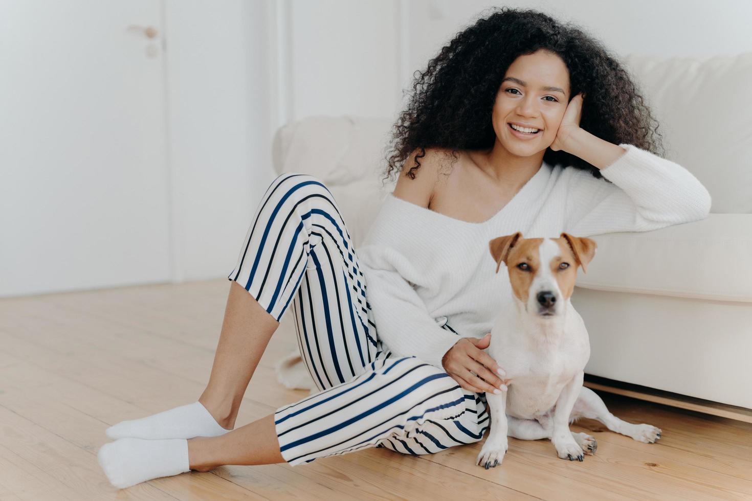 horizontale aufnahme einer schönen lockigen frau lehnt sich am sofa, sitzt auf dem boden in der nähe eines rassehundes, trägt einen pullover, gestreifte hose und socken. Afroamerikanerin posiert mit Haustier. menschen- und tierkonzept foto