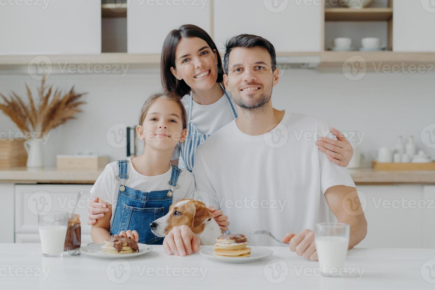 familie, freizeit, zeitvertreib, essenskonzept. vater, mutter und tochter, jack russell terrier hund posieren alle zusammen vor der kamera vor dem kücheninterieur, erfreuen sich daran, pfannkuchen zu essen und frische milch zu trinken foto