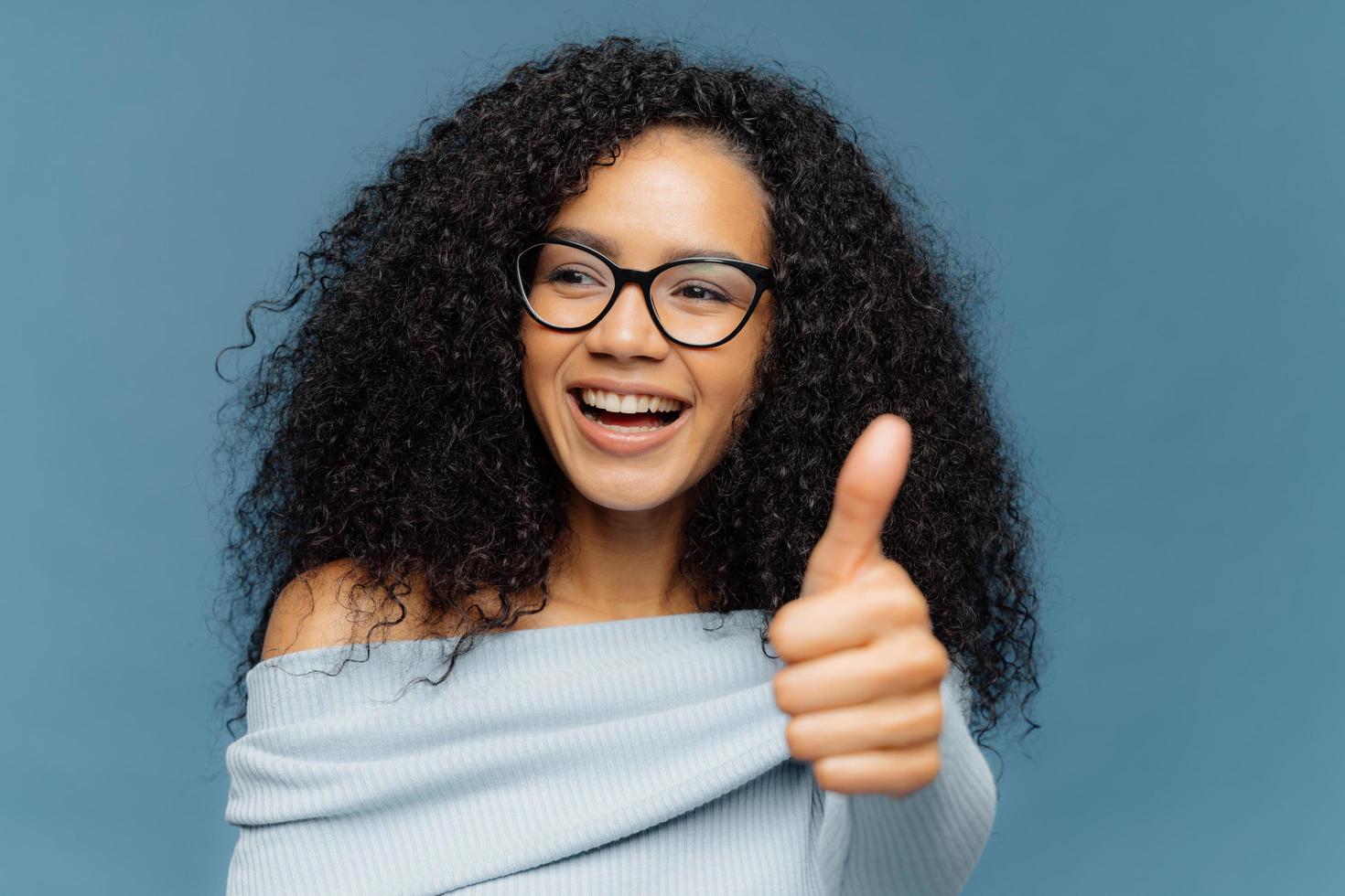 ich mag es porträt einer glücklichen frau mit afrofrisur, zeigt daumen nach oben, gibt eine positive meinung ab, schaut zur seite, trägt eine optische brille und einen pullover, isoliert auf blauem hintergrund. Menschen, Vereinbarung, Zustimmung foto