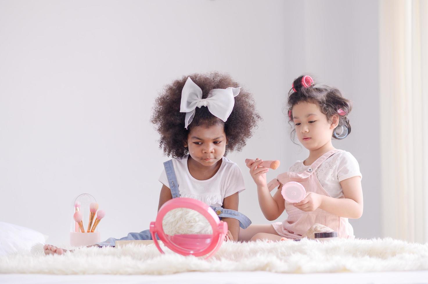 Ein süßes kleines asiatisches Mädchen und eine afrikanische Freundin tragen in ihrem Schlafzimmer glücklich Make-up-Pinsel mit Puder auf foto