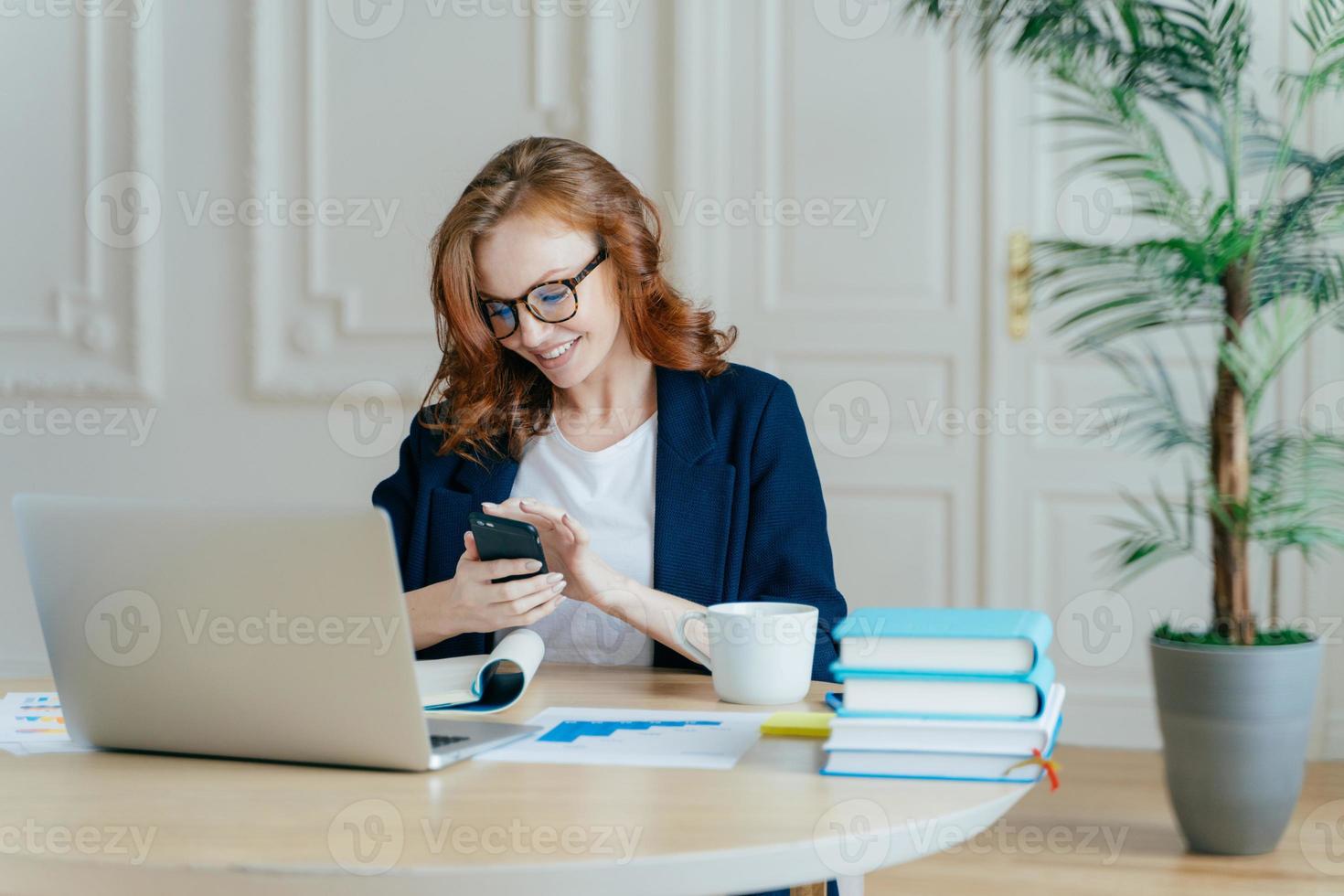 Technologie- und Kommunikationskonzept. Rothaarige junge Frau checkt E-Mail-Box auf modernem Handy, hat ein angenehmes Lächeln, studiert Dokumente, besitzt ein großes Unternehmen, benutzt einen Laptop-Computer für die Arbeit foto