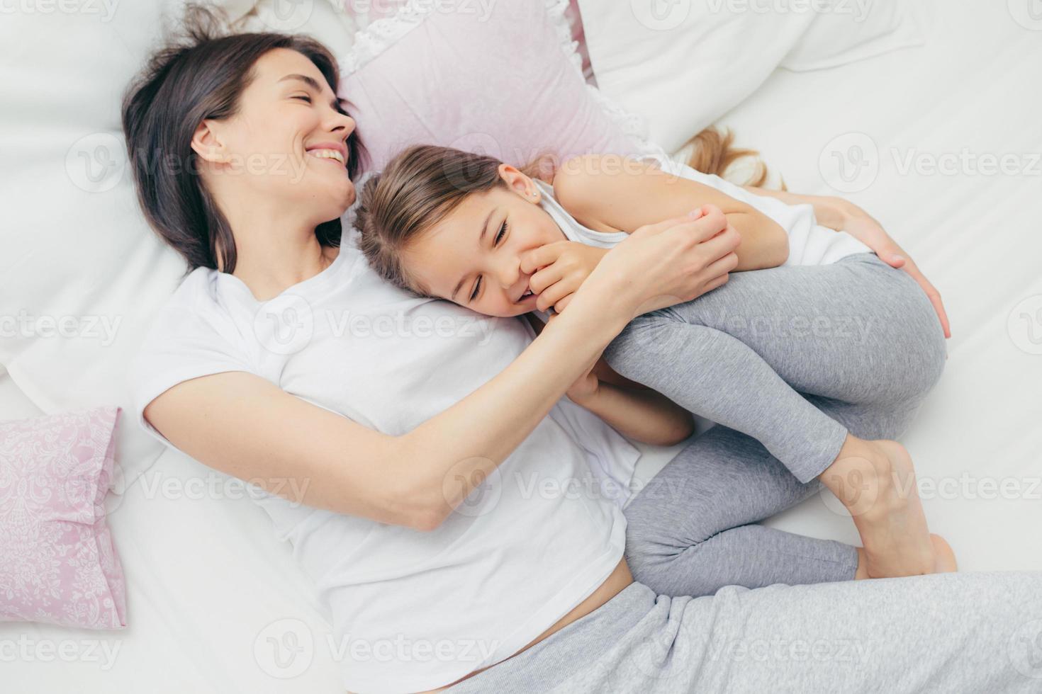 Entzücktes weibliches Kind kichert freudig, während es mit seiner Mutter in einem bequemen Bett spielt, positives Lächeln auf den Gesichtern hat, Schlafanzüge trägt, nach dem Aufwachen gute Laune hat. menschen, freizeit und schlafkonzept foto