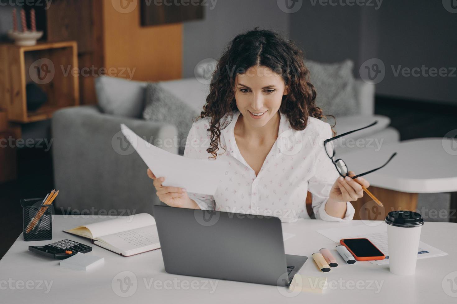 lächelnde, frohe italienische Frau, die zu Hause online Rechnungen bezahlt, während sie im Wohnzimmer sitzt foto