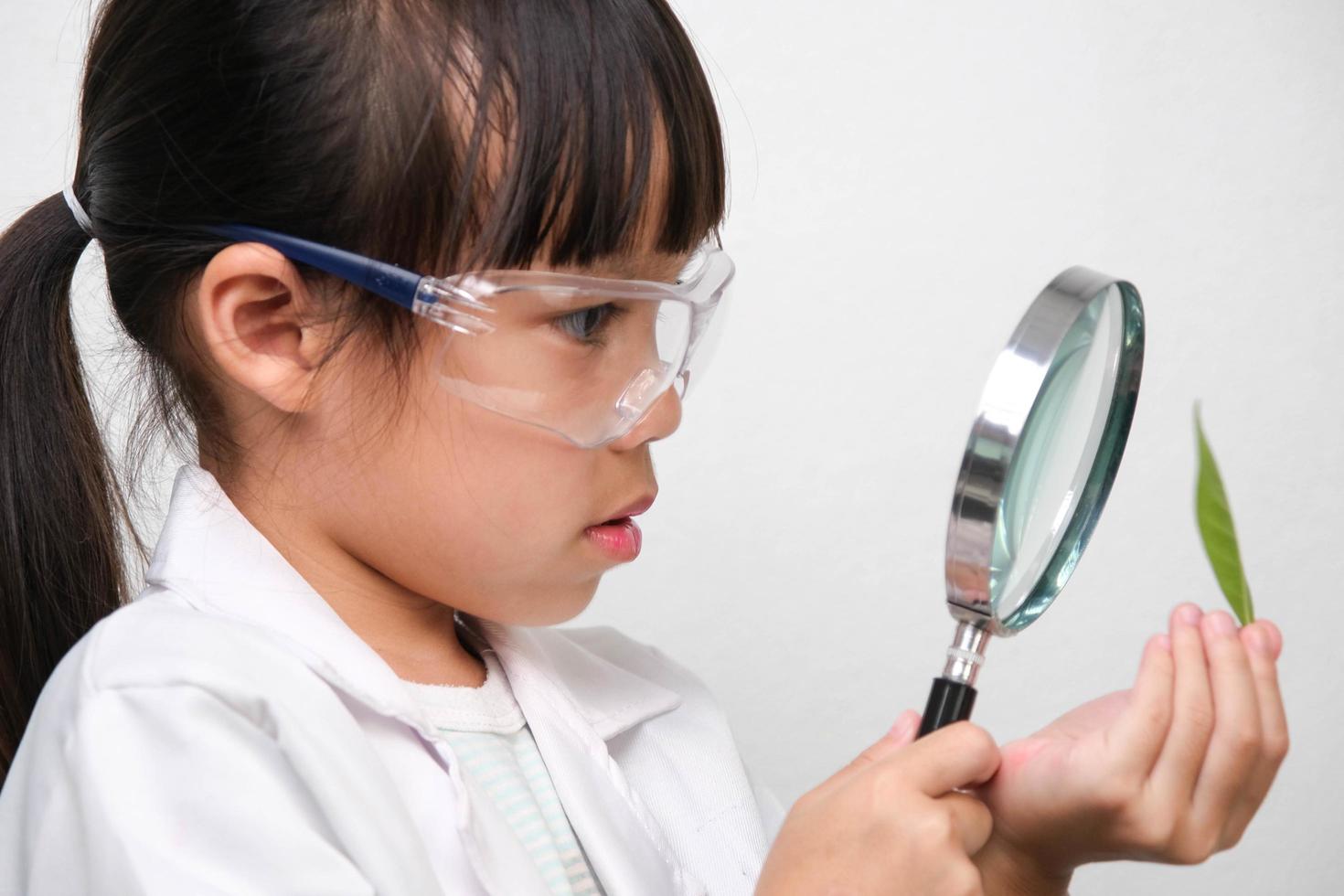 Porträt eines kleinen Mädchens mit Brille, das eine Lupe hält und Blätter in Forscher- oder Wissenschaftsuniform auf weißem Hintergrund betrachtet. kleiner Wissenschaftler. foto