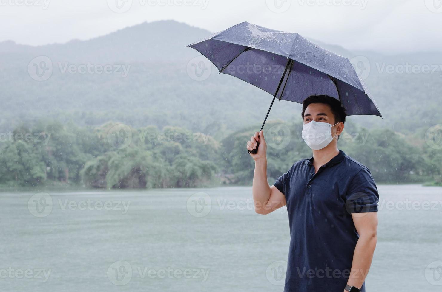 asiatischer mann, der gesichtsmaske trägt und regenschirm hält, um sich vor krank und nass zu schützen, wenn es in der monsunzeit regnet. foto