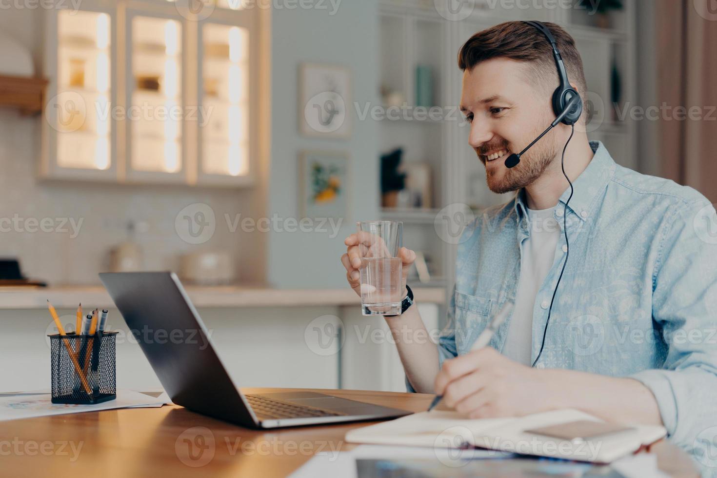 Fernlernkonzept. Millennial-Student im Headset beobachtet Webinar hat Online-Unterricht während der Quarantäne genießt Internet-Bildung trinkt Wasser aus Glas macht sich Notizen Posen am Desktop foto