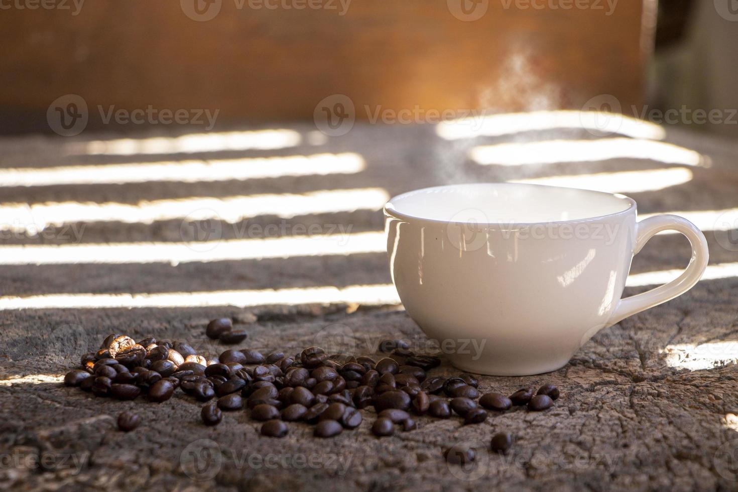 Weiße Kaffeetasse aus Keramik auf einem rustikalen Holztisch mit Kaffeebohnen unter der Morgensonne durch den Zaun. foto