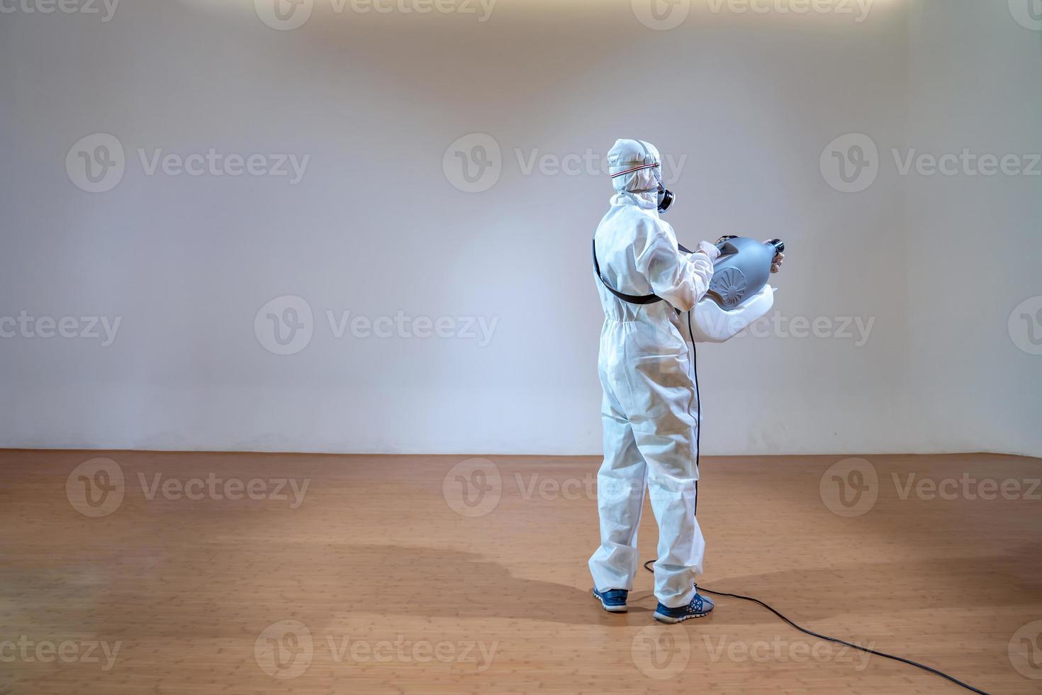 professioneller Techniker im Präventionsanzug sprüht Sterilisationslösung mit elektrischer Sprühmaschine auf Holzboden und weißem Hintergrund mit Studiolicht. foto