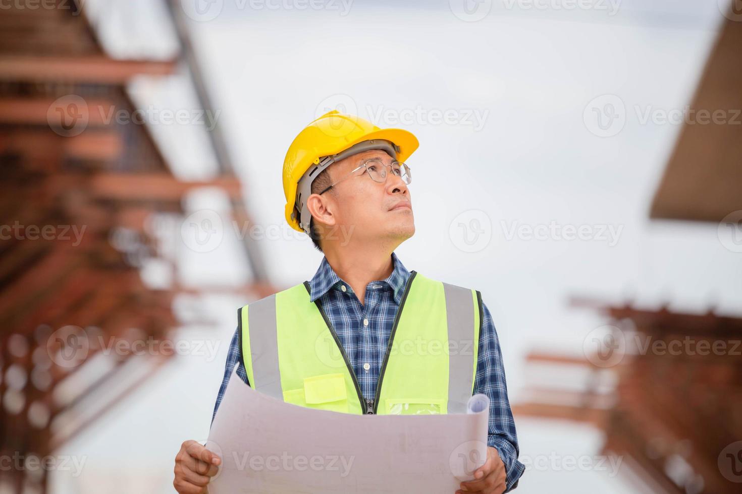 asiatischer bauingenieur, der projekt auf der baustelle überprüft, architekt mit bauplänen auf einer baustelle, vorarbeiter in helm auf der infrastrukturbaustelle foto