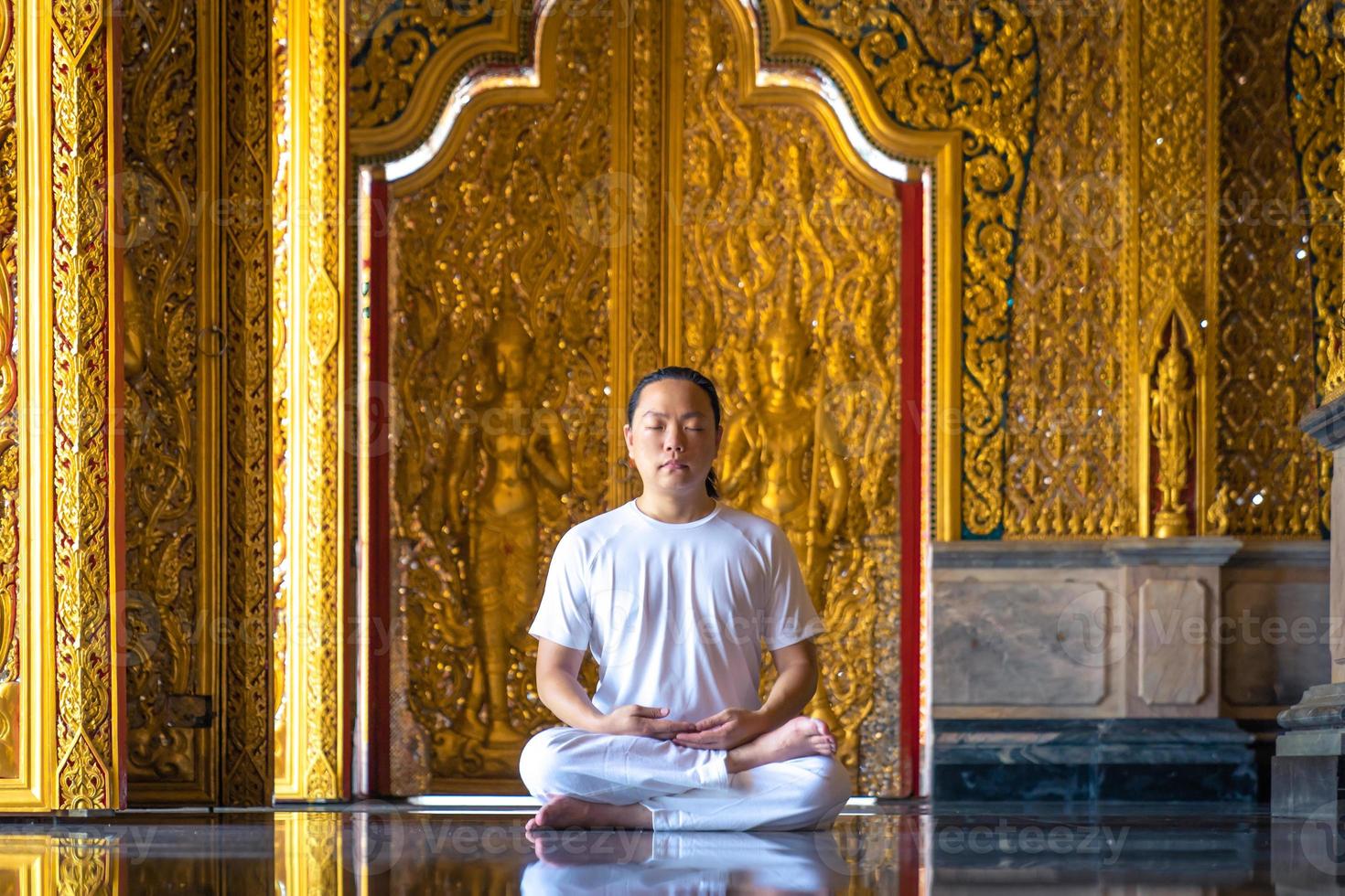 asiatischer langhaarmann entspannt meditation mit ganz weißem kostüm sitzt vor der goldenen tapete des buddisten im tempel, thailand. foto